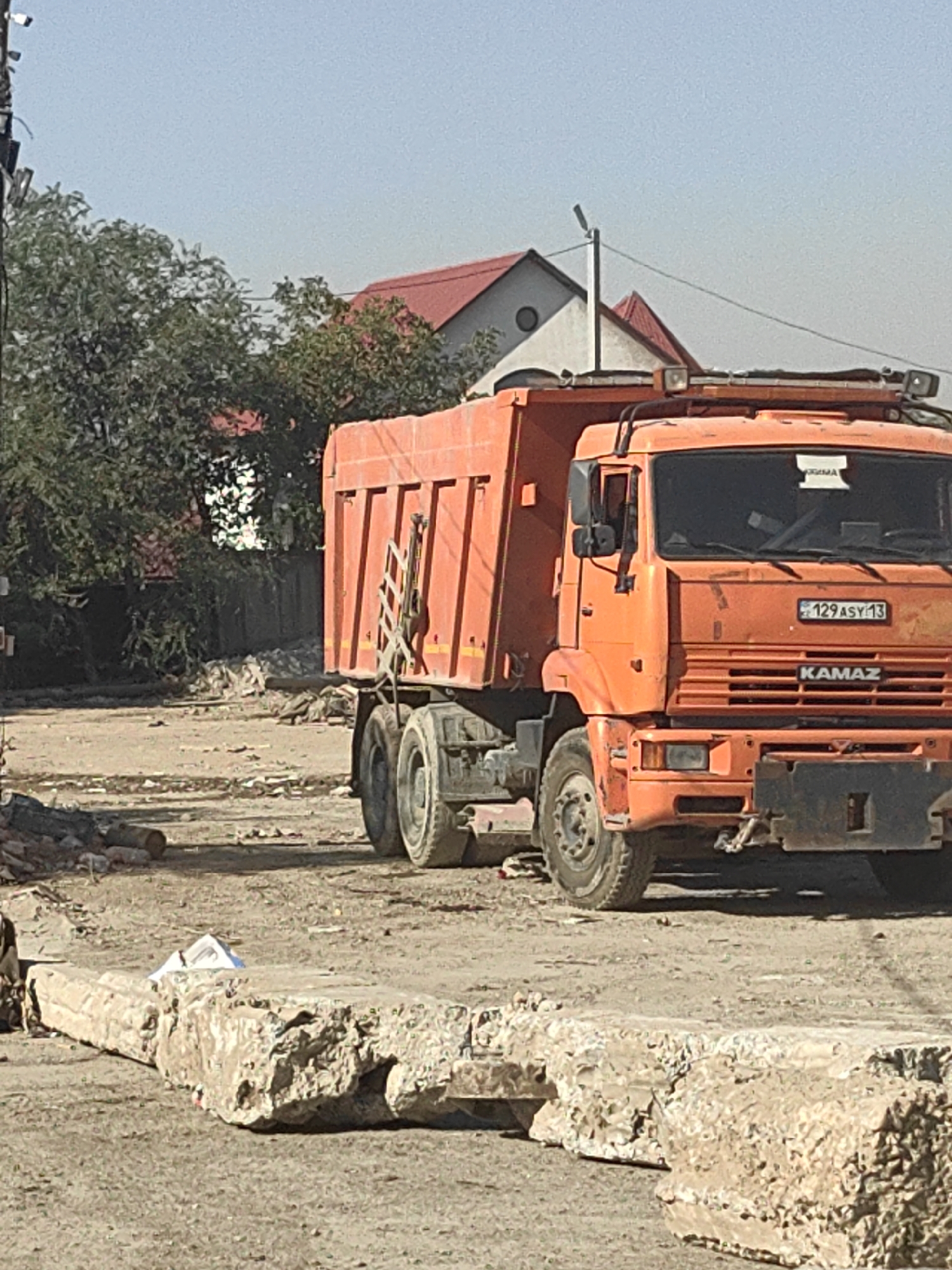 An orange Kamaz dump truck is parked on a dirt road in a construction or demolition site. The truck has a license plate that reads "129 ASY 13". In the foreground, there are large concrete debris. In the background, there are trees and houses with red roofs. The sky is clear and blue. The scene appears to be in Shymkent, Kazakhstan, as indicated by the license plate format.