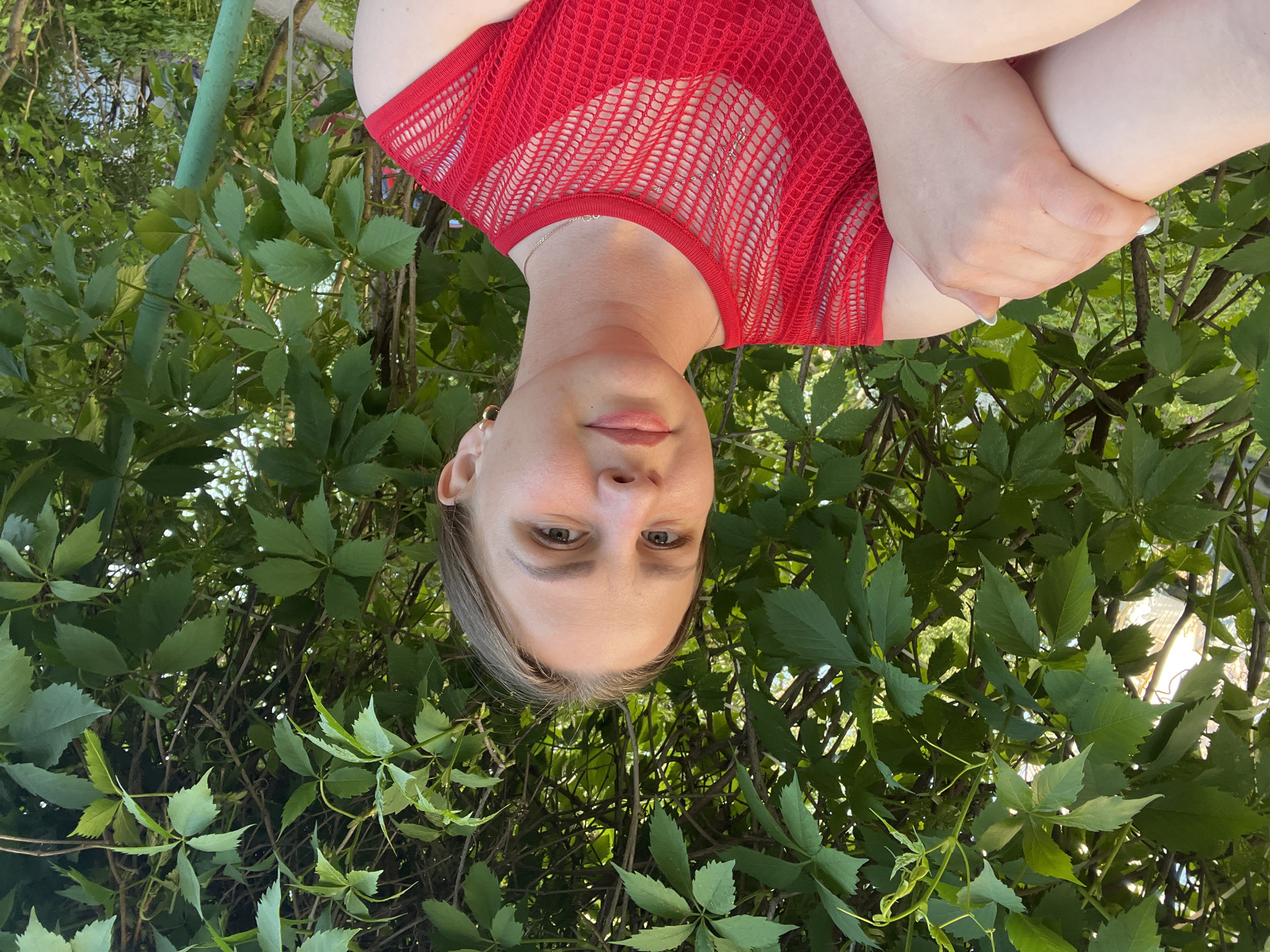 A woman wearing a red mesh top is positioned in front of dense green foliage. Her arms are crossed in front of her. The location is Izhevsk, Russia, based on contextual clues not visible in the image.