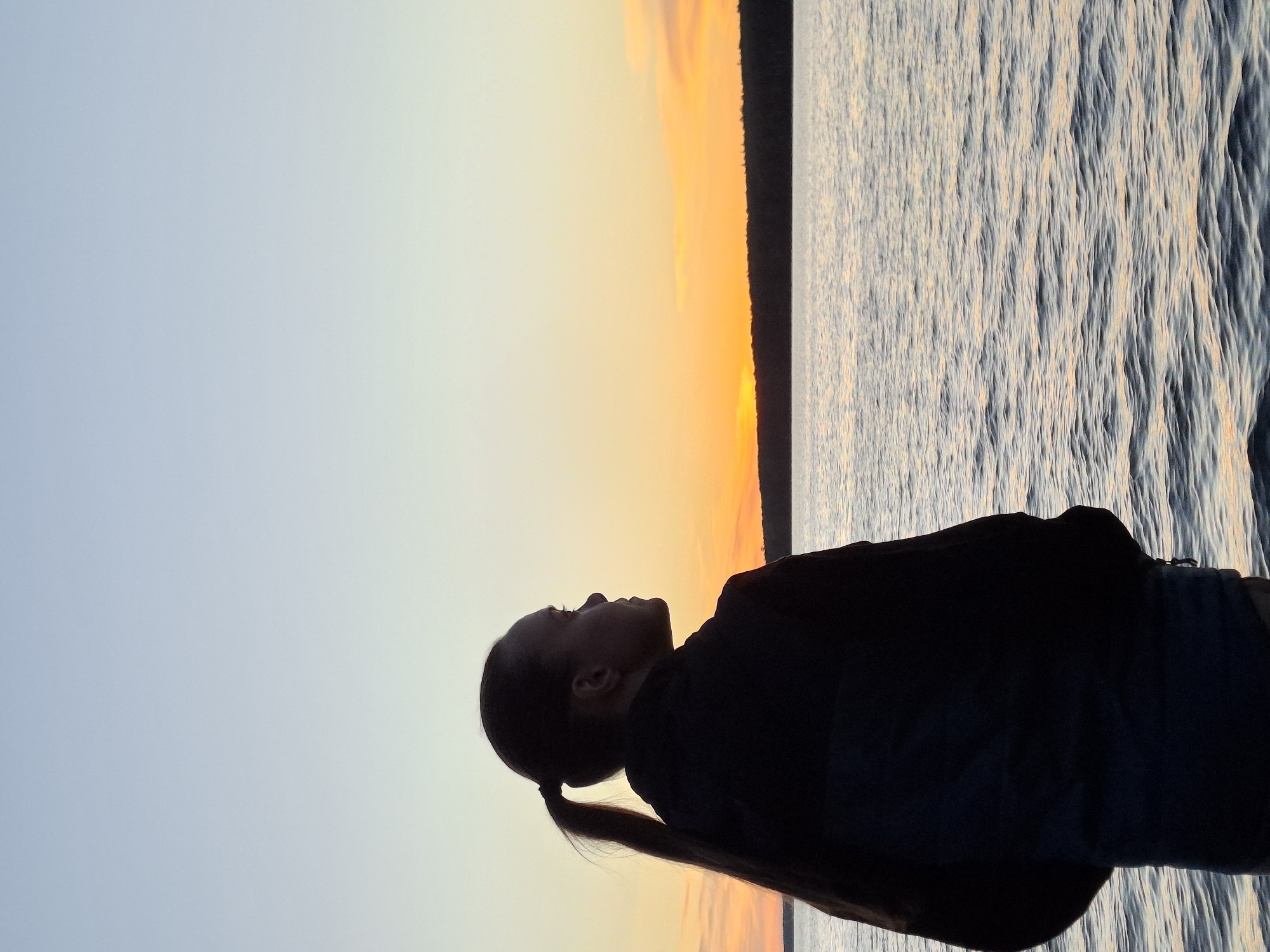 The image shows a silhouette of a young woman from the shoulders up, facing right. She has her hair in a ponytail. She is wearing a dark jacket with a blue section on the lower back. She is standing on a body of water, possibly a river or lake, with a tree line visible on the horizon. The sky is clear with soft pastel colors indicative of a sunset or sunrise. The water has small ripples. The setting appears to be a natural landscape, likely on the outskirts of Chaikovsky, Russia, given the time of day and the presence of a large body of water. The overall mood is peaceful and contemplative.