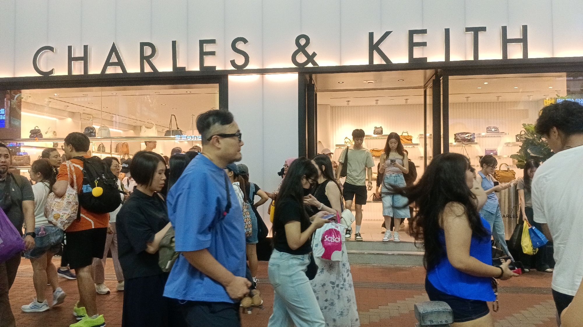 The image shows a street scene in Hong Kong, China, in front of a Charles & Keith store. The store's name is prominently displayed in large, illuminated lettering above the entrance. Inside the store, handbags are displayed on shelves. A group of people are walking on the sidewalk in front of the store, with some looking towards the store and others engaged in conversation. The sidewalk appears to be paved with cobblestones or bricks. The overall lighting suggests it is daytime, possibly late afternoon.