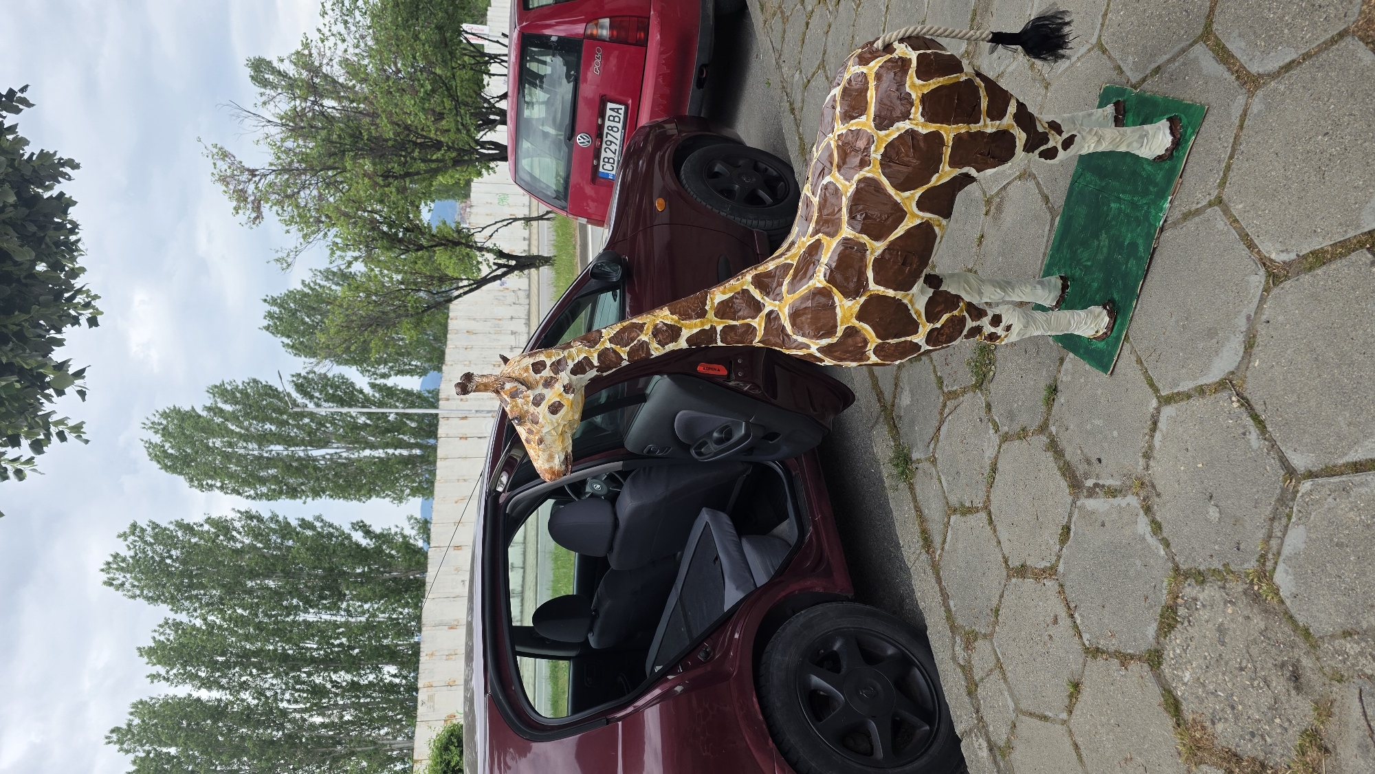 A life-size giraffe sculpture made of painted paper mache and other materials is partially placed inside a dark red car with its door open. The giraffe's tail is black, and its body is brown with yellow spots. The giraffe stands on a small green platform on a grey, hexagonal-patterned pavement. The car's license plate is visible: CB 2978 BA. Behind the car is a red Volkswagen Polo, and in the background are green trees against a cloudy sky. The scene is located on a street in Blagoevgrad, Bulgaria.