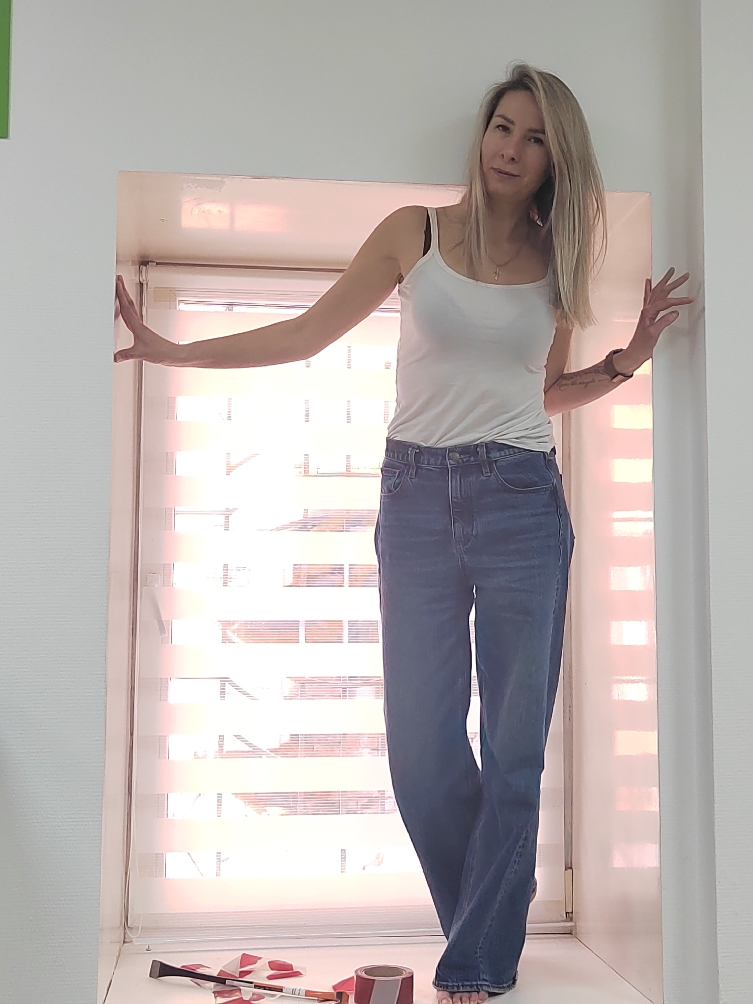 A woman is standing in a window alcove. She is wearing a white tank top and blue jeans. She has blonde hair. Her arms are extended, and her hands are touching the sides of the alcove. There is a window behind her with horizontal blinds. On the window sill below are a utility knife, an X-ACTO knife and a roll of tape. The scene appears to be indoors. There is no information to confirm that the location is in Votkinsk, Russia.
