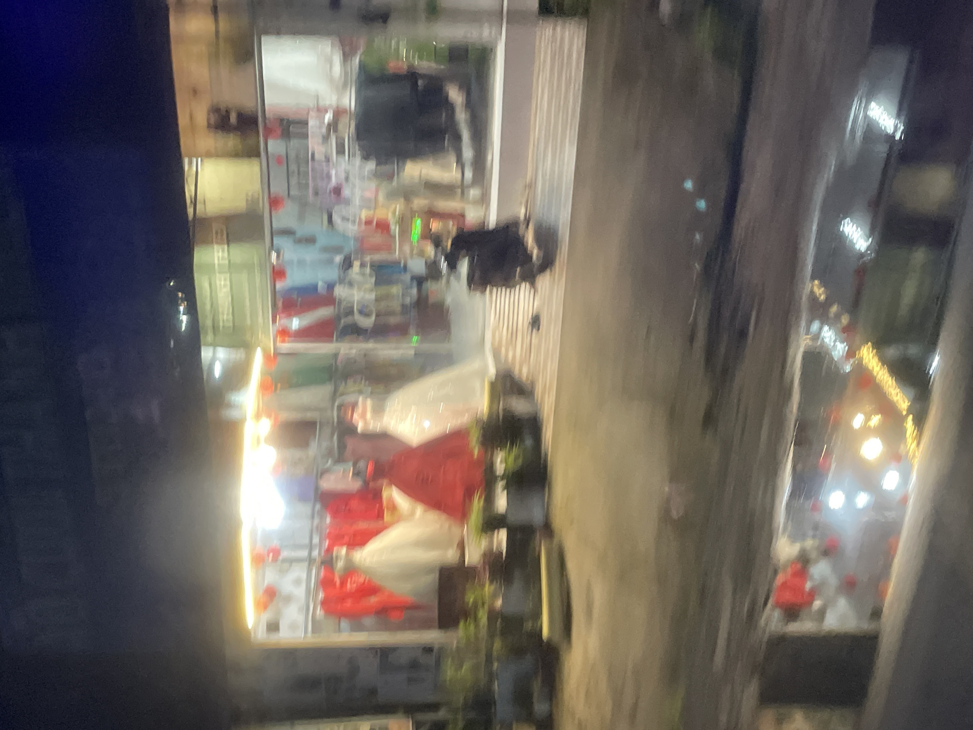 The image shows a street scene at night in Phú Khánh, Vietnam. The perspective is slightly tilted, giving the impression of looking down at an angle. On the left, a brightly lit storefront displays several formal gowns, appearing to be wedding dresses or traditional Vietnamese attire like Ao Dais. The dresses are in white and red, with some intricate detailing visible. The storefront's interior also shows shelves stocked with various items, suggesting a retail establishment.
In the foreground, a dark, shadowy area dominates the left side of the frame, potentially a wall or a building facade. To the right of the storefront, a paved walkway or street is visible. Reflections on the wet pavement suggest it may have recently rained or is damp. Further to the right, another storefront or building is seen, also illuminated with lights, with what appear to be more displays or objects inside. Decorative red lanterns are visible hanging from the ceiling or structures in the distance, hinting at a festive occasion or a general display of cultural elements.
The overall atmosphere is that of a commercial street at night, with shops open and lights on. The presence of formal gowns and decorative lanterns implies a cultural context possibly related to celebrations or formal events. The lighting is a mix of bright shop lights and the softer glow of streetlights or decorative illuminations. No people are clearly identifiable as subjects in the scene, though there might be indistinct figures within the shops or on the street, blurred by the low light and motion. No text is legible in the image.