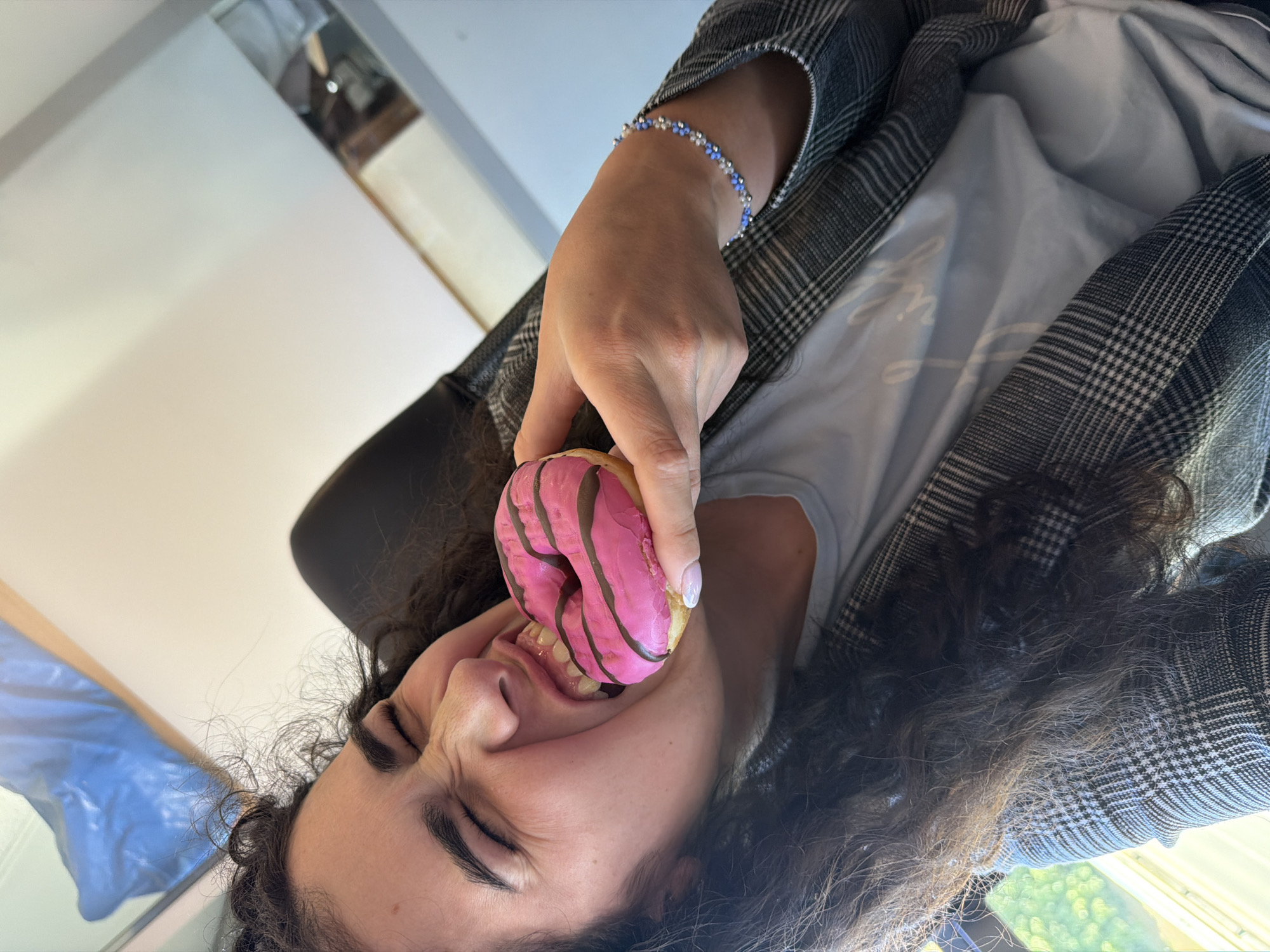 A young woman with dark curly hair is taking a bite out of a pink frosted donut with dark chocolate stripes. She is wearing a gray plaid blazer over a light blue t-shirt and a beaded bracelet on her left wrist. Her eyes are closed, and her mouth is open wide to accommodate the donut. The background is blurred, but appears to be an indoor setting with natural light coming from a window. It is not possible to determine the location in Sofia, Bulgaria.