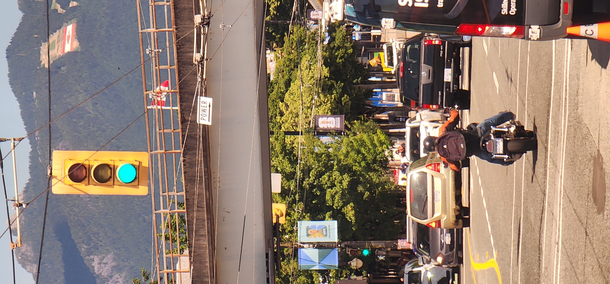 The image shows a street scene in a city with a prominent traffic light displaying green. A motorcycle with a rider wearing a backpack is in the foreground, stopped at the intersection. Several cars are also visible in traffic. In the background, a large Canadian flag is displayed on a hillside, and banners hang from poles above the street. A sign with the word "POWER" and an arrow pointing to the right is attached to an overpass. The scene appears to be on a sunny day with trees and mountains in the distance.