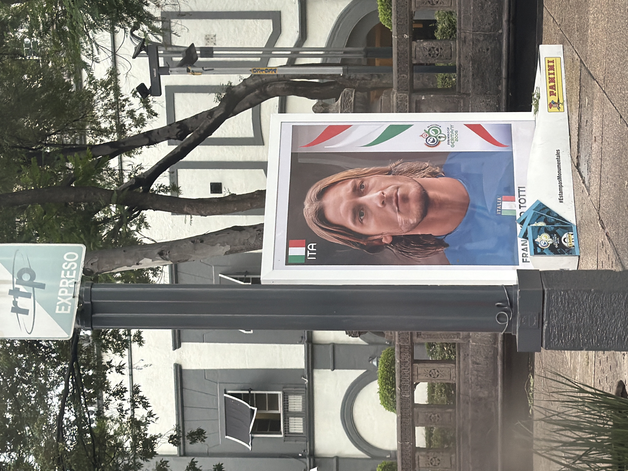 The image shows a poster attached to a lamppost, likely in Mexico City, Mexico. The poster features a portrait of Italian soccer player Francesco Totti, referencing Italy's victory in the 2006 FIFA World Cup. There is also signage for "rtp EXPRESO" on the lamppost. Other details include a sticker for the Panini sticker album collection, indicating a focus on soccer memorabilia. The background features a light gray two-story building with dark gray trim, trees, and a stone structure behind the poster.