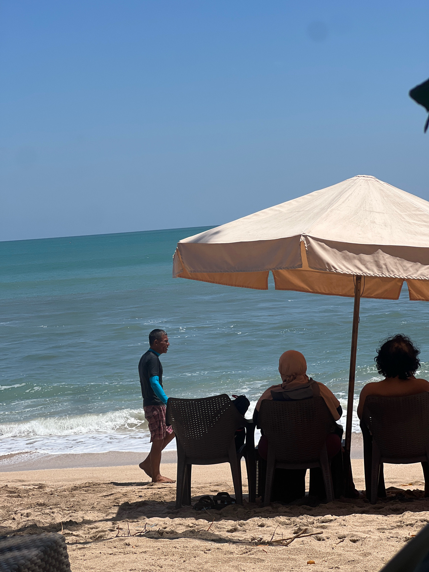 The image depicts a beach scene in Kuta, Indonesia, on a clear, sunny day. In the foreground, two individuals are seated in chairs, facing the ocean and partially obscured by a large, beige beach umbrella. The person on the left is wearing a headscarf. To the left of the seated figures, a man walks along the shore, wearing a dark t-shirt, light-colored shorts with a plaid pattern, and blue arm coverings. He is facing away from the viewer and towards the water. The ocean is calm with gentle waves rolling onto the sandy beach. The sand is light brown and shows some scattered debris. The sky is a clear, bright blue.