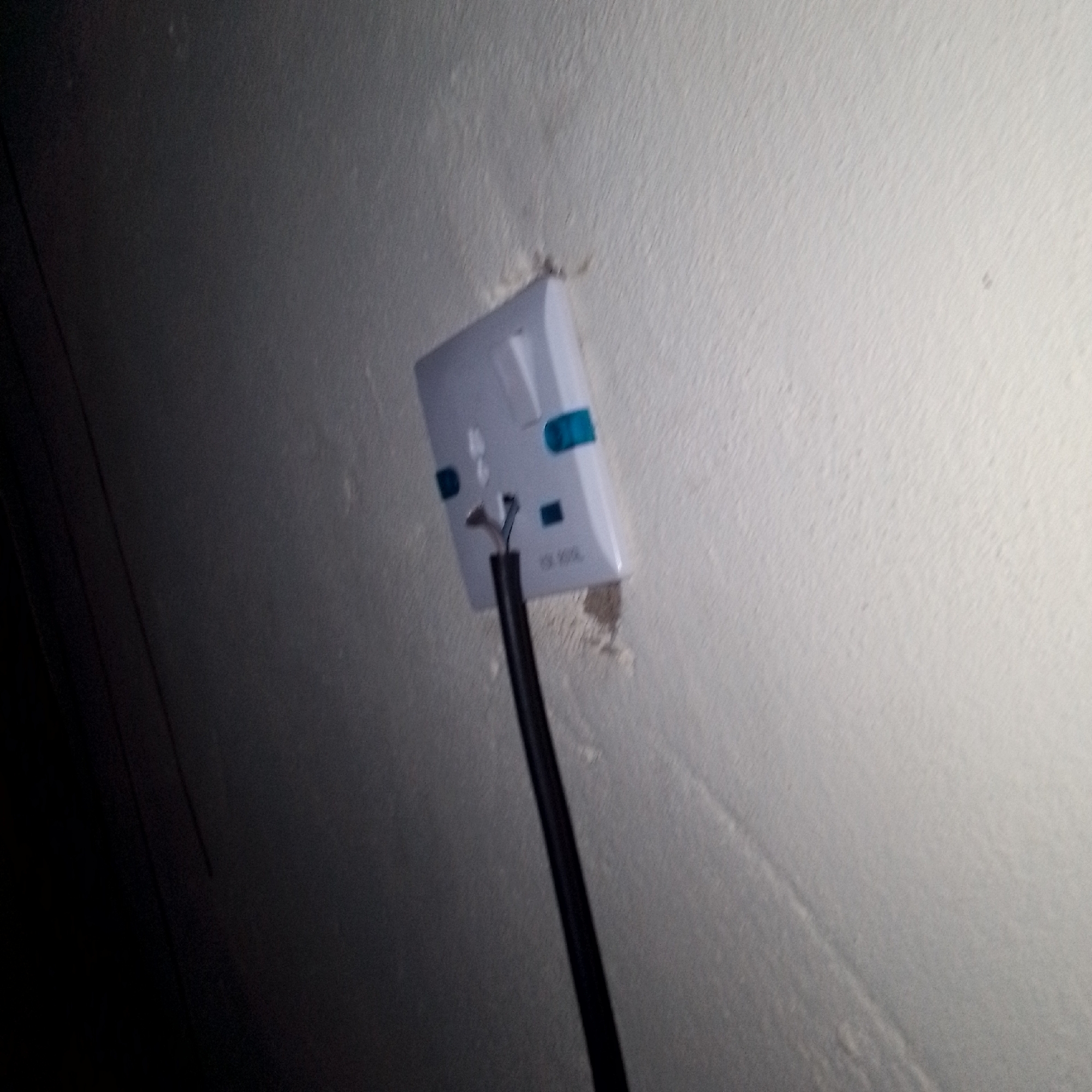 The image shows a white electrical outlet mounted on a textured, light-colored wall. A black electrical cord extends from the bottom of the outlet, with its wires exposed at the connection point. The outlet has blue accents on its face. The surrounding wall shows some signs of wear or damage around the outlet. The scene is dimly lit, with a dark area on the left side of the frame.