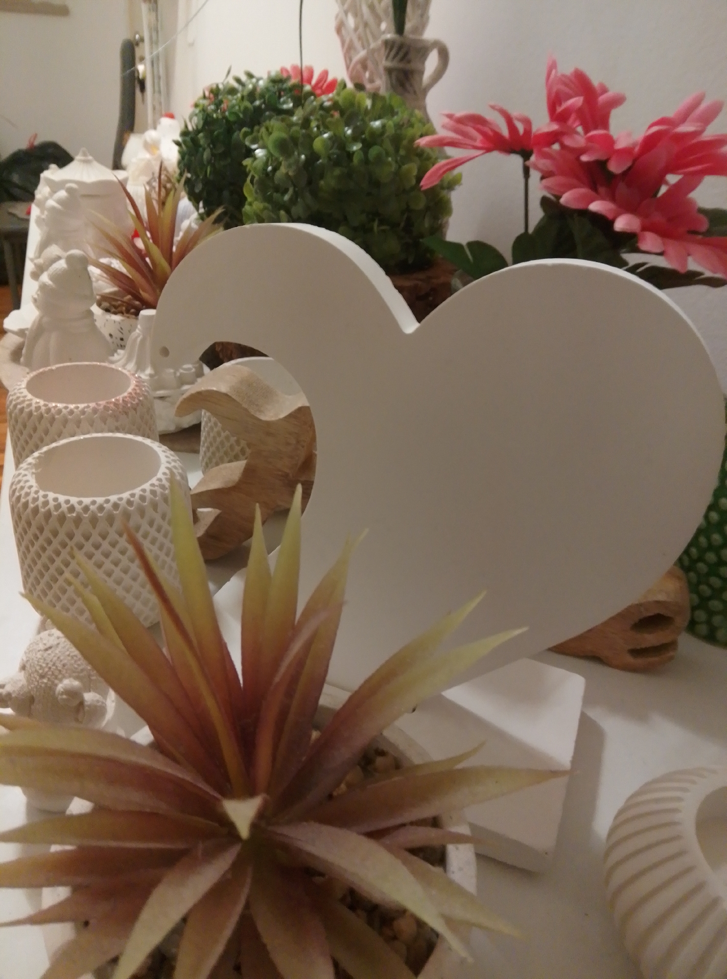 The image depicts a collection of decorative items on a white surface, possibly a shelf or table. In the foreground, a plant with long, spiky, yellowish-red leaves is planted in a white, textured pot. To the left of the plant are two more white, textured cylindrical containers. Behind these, a white, decorative figurine of a rounded figure wearing a hat is visible. Further back and to the left, a white, intricate lidded container sits next to a leafy green artificial plant. To the right of the green plant, a cluster of bright pink artificial flowers is arranged. Dominating the center of the image is a large, white, heart-shaped cutout. Behind the heart, a wooden sculpture with a curved shape is partially visible, as is another artificial plant with green, rounded leaves, and a textured green object with circular indentations. The background is largely out of focus, showing hints of white walls and possibly other objects. There are no people visible in the image, and no actions or interactions are depicted. The lighting appears to be artificial, casting soft shadows. The scene is likely set indoors in a domestic or retail environment. The city of Wedel, Germany is not mentioned in the image or implied by its contents.