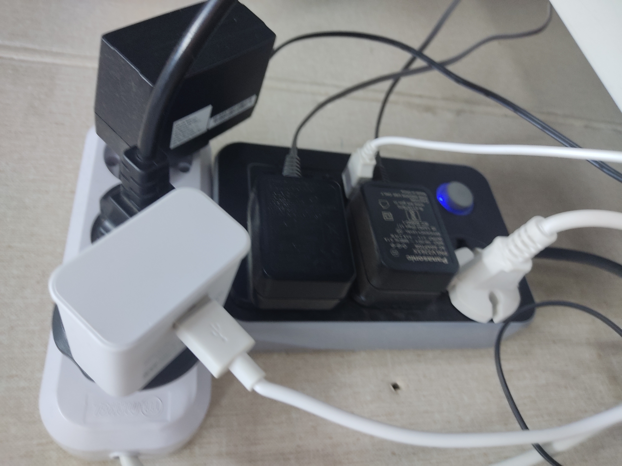 A white power strip with multiple adapters is plugged into a wall socket. A white cable is connected to one of the adapters, and a black cable is connected to another. Two black power adapters are plugged into the power strip. One adapter has a blue light indicator. The wall appears to be a textured, light-colored surface. The scene is indoors, in an indoor setting.