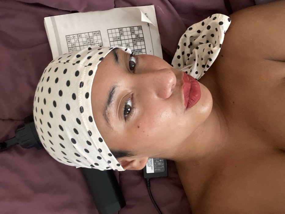 A woman with light brown skin wears a white and black polka dot headscarf. She lies on a purple bed with a partly completed Sudoku puzzle near her head. A black massage gun and its adapter rest close by. Her lips are colored a matte red. She gazes into the distance with a slightly contemplative expression.