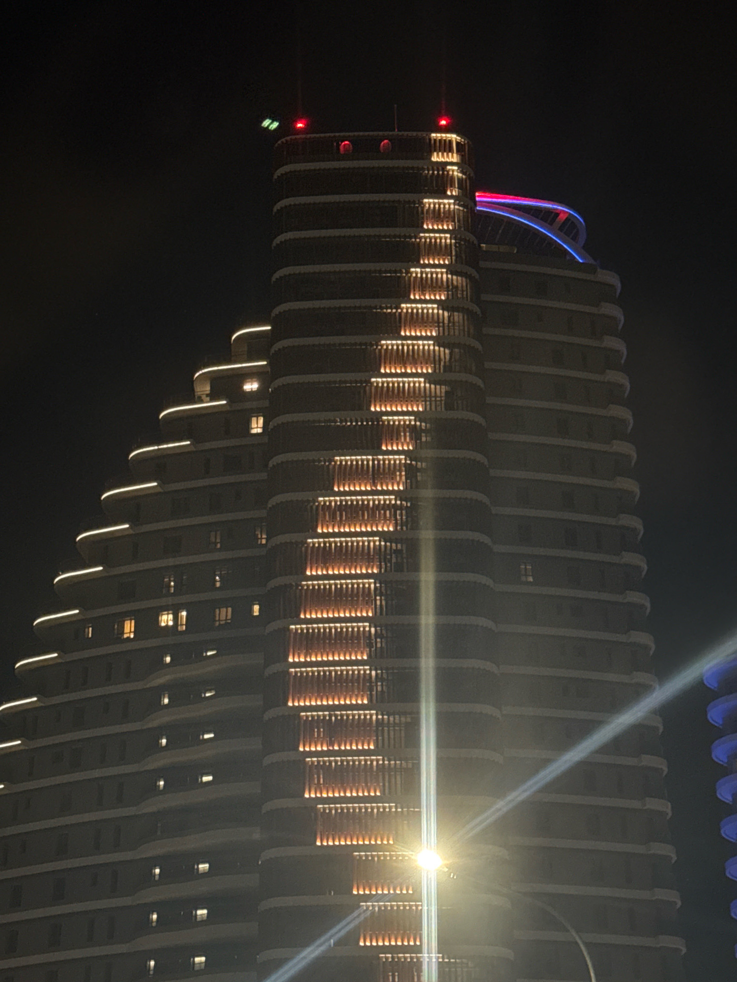 The image shows a modern, multi-story building at night in Germasogeia Municipality, Cyprus. The building has a stepped, terraced design with curved edges. It is illuminated by various lights, including white lights along the edges of each tier, warm orange lights in vertical panels on one side, and a red light at the very top. A vibrant blue and red neon-like structure curves over the upper section of the building. The sky is dark, indicating nighttime. A bright light source, possibly a streetlamp, creates a strong glare across the lower right portion of the image, with a beam of light extending upwards. To the right, another building with blue curved elements is partially visible. No people are discernable in the image.