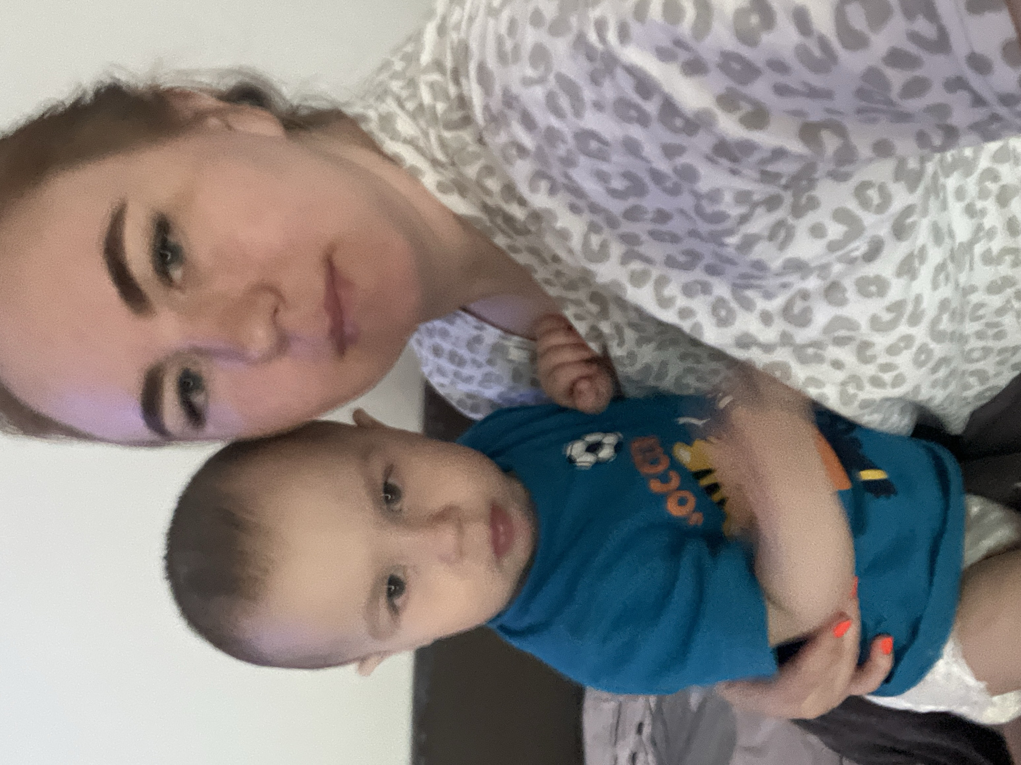 The image shows a woman with light skin and dark eyebrows, wearing a gray and white patterned shirt, holding a baby. The baby, also light-skinned, is wearing a blue shirt with "soccer" written on it and has a white diaper. The location is in Chaikovsky, Russia.