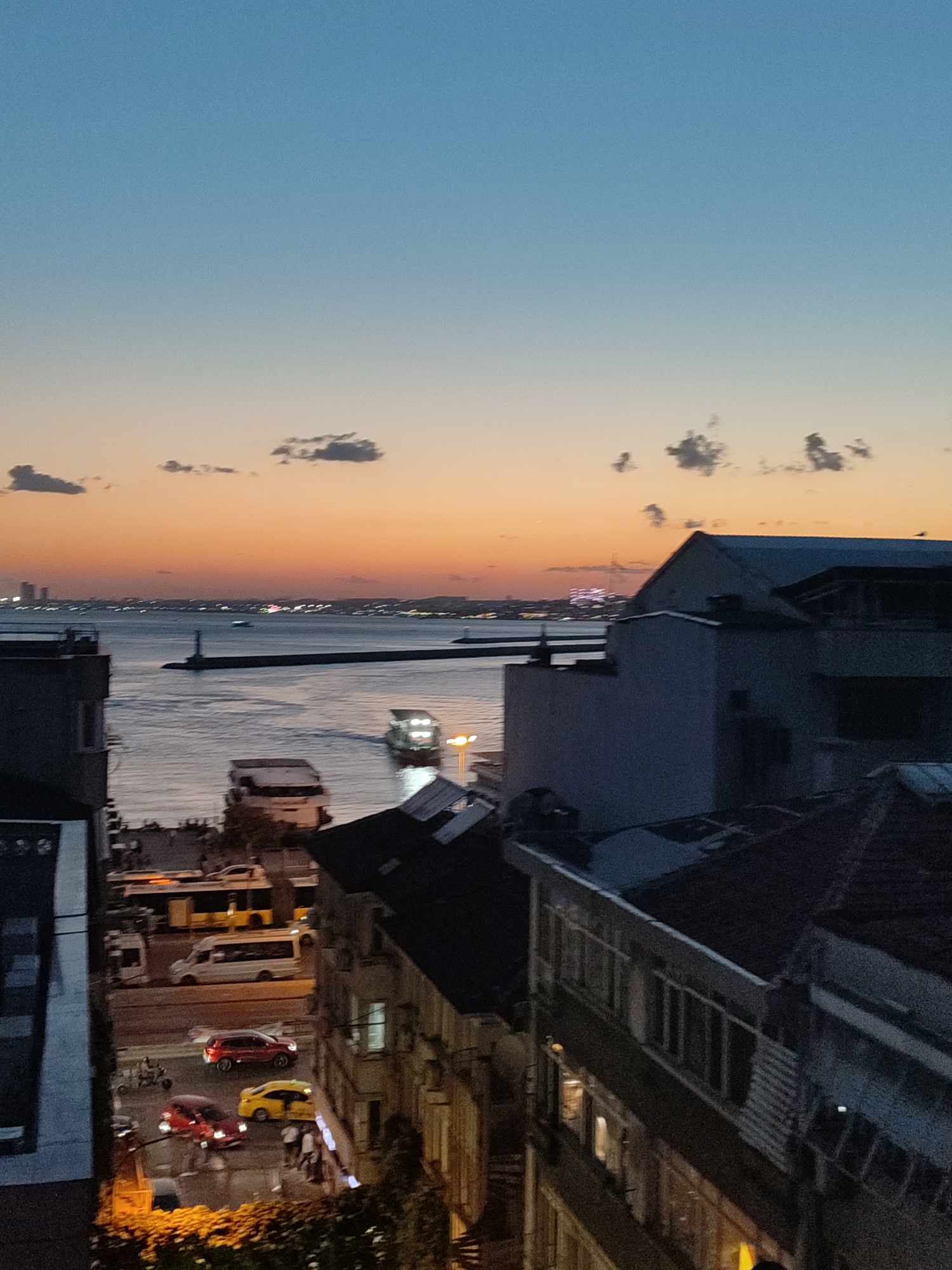 The image depicts a twilight scene overlooking the city of Kadıköy, Turkey, with the Bosphorus strait in the foreground. The sky displays a gradient of deep blue at the top transitioning to a warm orange and pink hue near the horizon, indicative of sunset. Several small, scattered clouds are visible against the colorful sky.
The Bosphorus strait is calm, reflecting the ambient light. A ferry or boat is navigating the water, its lights illuminated. A lighthouse stands on a pier extending into the water. On the distant cityscape, buildings and lights are visible, suggesting a densely populated urban area.
In the mid-ground and foreground, buildings of varying architectural styles are present. Some structures appear to be older, with visible windows and balconies, while others are more modern. On the street level, several vehicles, including cars and a van, are visible. Some cars are red and yellow. Pedestrians are also present on the sidewalks. The overall atmosphere suggests a lively urban environment transitioning into evening. The image is taken from an elevated perspective, likely a balcony or higher floor of a building.