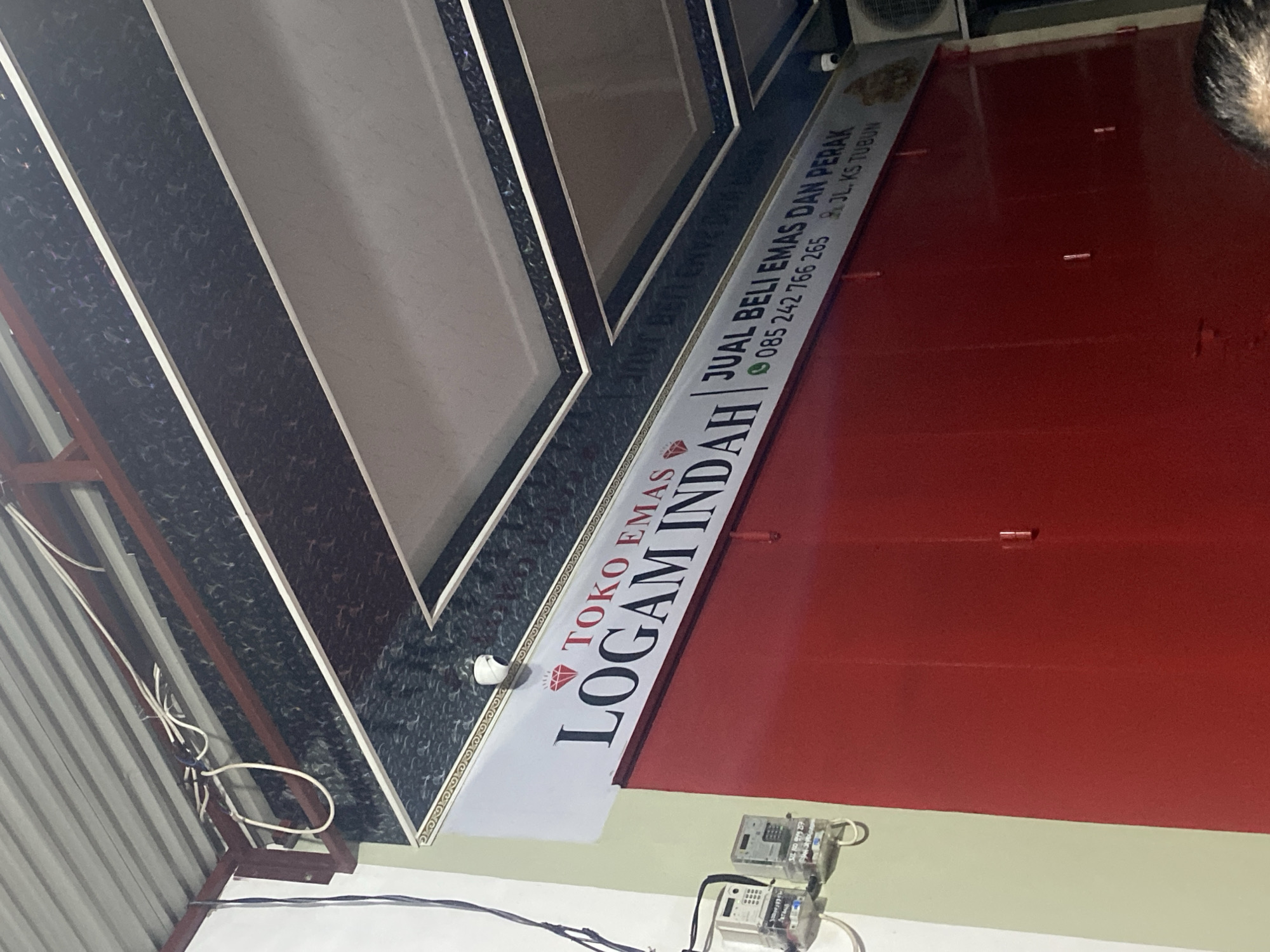 A sign is mounted on a red wall in front of a building. The sign reads "TOKO EMAS LOGAM INDAH JUAL BELI EMAS DAN PERAK 085 242 766 265 JL. KS TUBUN". There are two electric meters attached to the wall to the left of the sign. The ceiling above the sign is decorated with a patterned material and recessed lighting. Wires are visible near the ceiling. A security camera is mounted above the sign. The overall environment appears to be a storefront or shop.