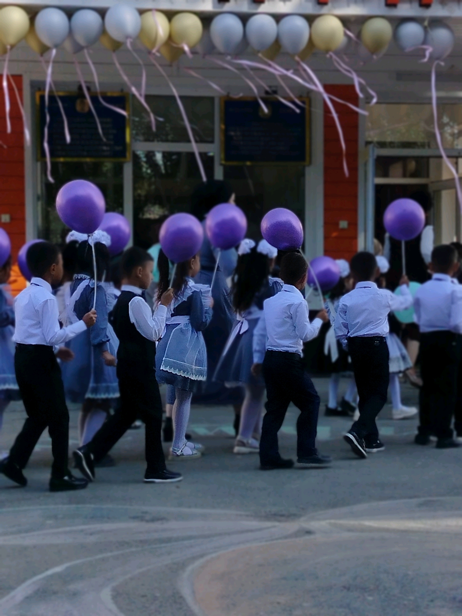 In the city of Kölkent, Kazakhstan, a group of school children is seen in a procession, each holding a purple balloon. The children are dressed in formal attire, with boys wearing white shirts and black pants, and girls in blue dresses with white collars and aprons. The balloons, tied with pink ribbons, float upwards, creating a festive atmosphere. The background shows a building with orange and red accents, possibly a school, with windows and doors indicating an indoor setting. The ground is paved, with some visible markings. The scene appears to be an outdoor school event, perhaps a celebration or a commencement ceremony.