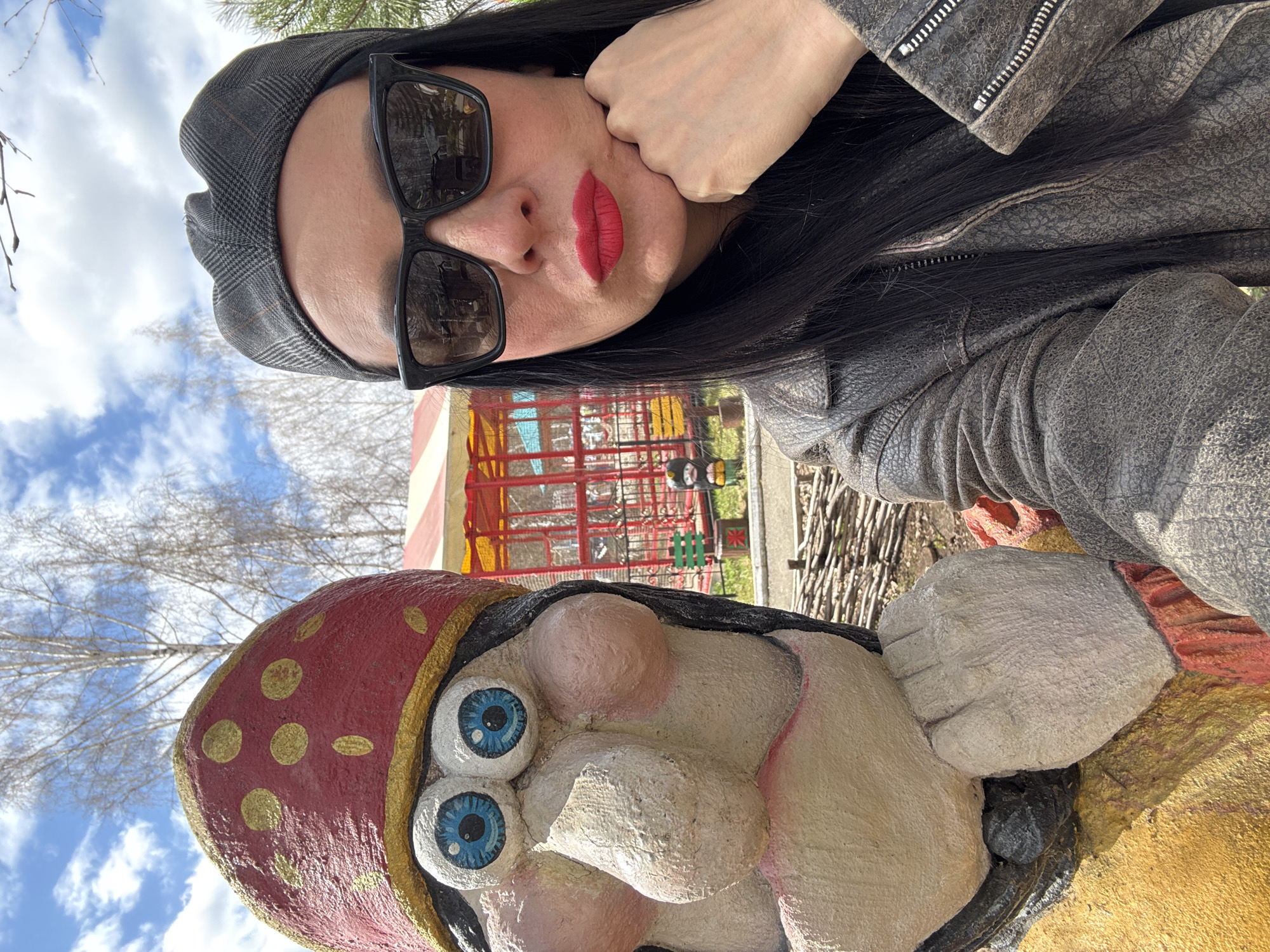 A selfie taken outdoors features a woman wearing a grey cap, black sunglasses, a grey leather jacket, and red lipstick. She rests her cheek on her hand. Below and slightly in front of her is a painted statue of a character wearing a red hat with gold polka dots. A playground with red and white striped elements is visible in the background. The location is Chaikovsky, Russia.