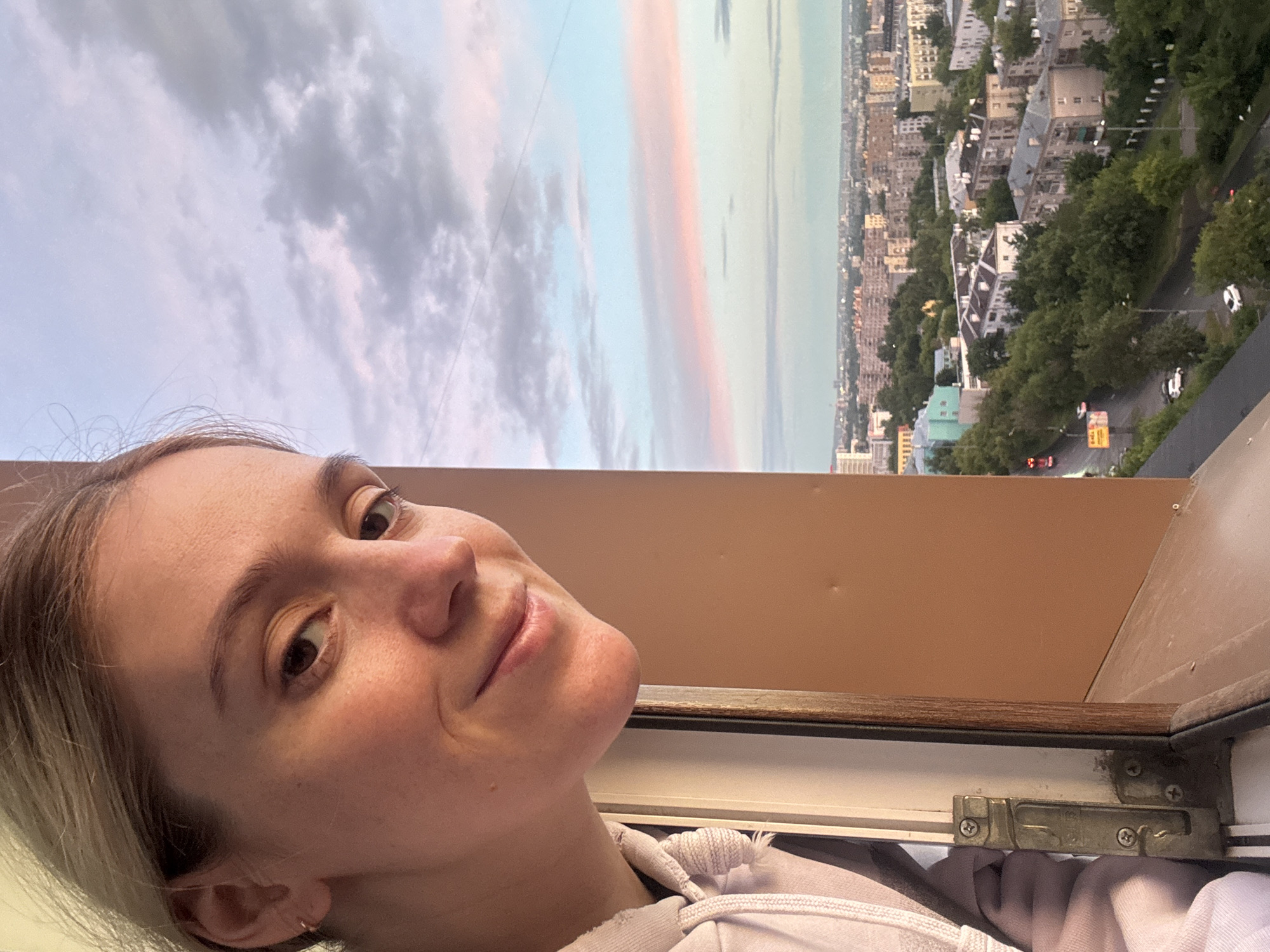 A young woman with blonde hair and fair skin leans out of an apartment window, looking towards the camera with a slight smile. Her face, partially obscured by the window frame, is illuminated by soft, natural light. She is wearing a light-colored hoodie.
Beyond the window, a panoramic view of a city unfolds under a twilight sky. The sky is painted with soft hues of pink, orange, and blue, with scattered clouds. Below, a dense urban landscape with numerous apartment buildings, some with a distinct Soviet-era architectural style, stretches to the horizon. Green trees line the streets and hills, interspersed with residential blocks. A road with a few cars is visible in the mid-ground, suggesting activity within the city. The image captures a serene moment of a person enjoying the cityscape from their dwelling. The city appears to be Nizhny Novgorod, Russia, judging by the architectural styles and the general urban layout.