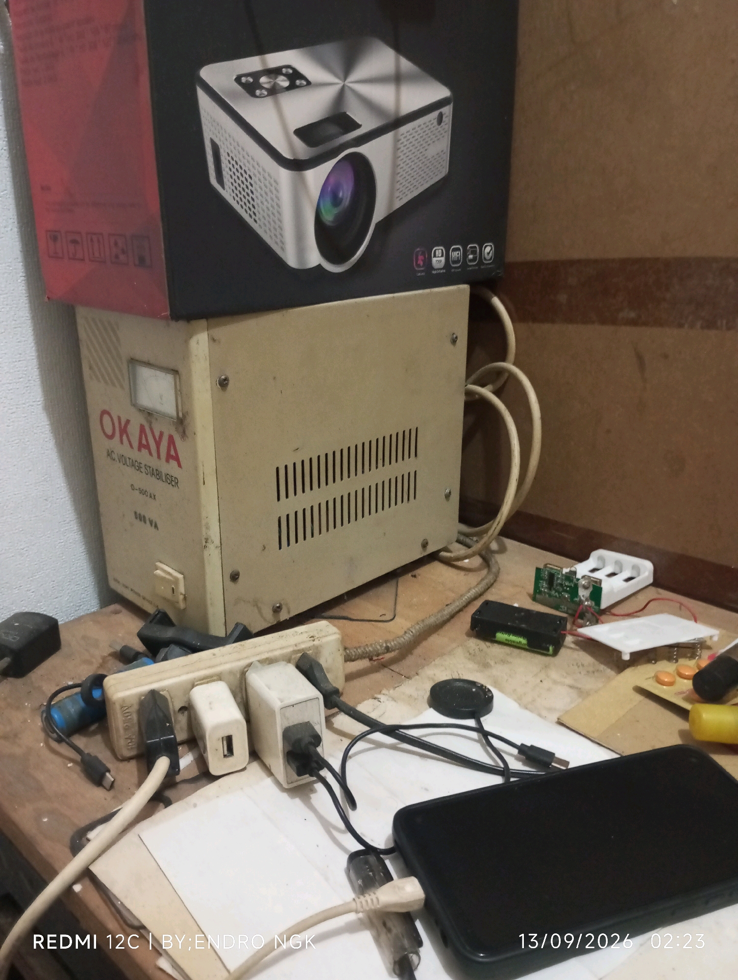A dimly lit indoor scene, likely a workshop or storage area, is depicted. In the foreground, a black smartphone lies on a wooden surface, partially obscured by charging cables connected to a power strip. The power strip itself is plugged into an AC voltage stabilizer unit, branded "OKAYA." Above the stabilizer, a projector is visible within its packaging. The background shows a textured wall, possibly unfinished wood or concrete. The overall impression is one of a functional, albeit cluttered, workspace. The timestamp "13/09/2026 02:23" and the text "REDMI 12C | BY:ENDRO NGK" are overlaid on the image, suggesting it was taken by Endro Ngk with a Redmi 12C phone on September 13, 2026, at 2:23 AM. There are no people visible in the image, and no specific location within Ringinanom, Indonesia, can be identified from the visual cues.