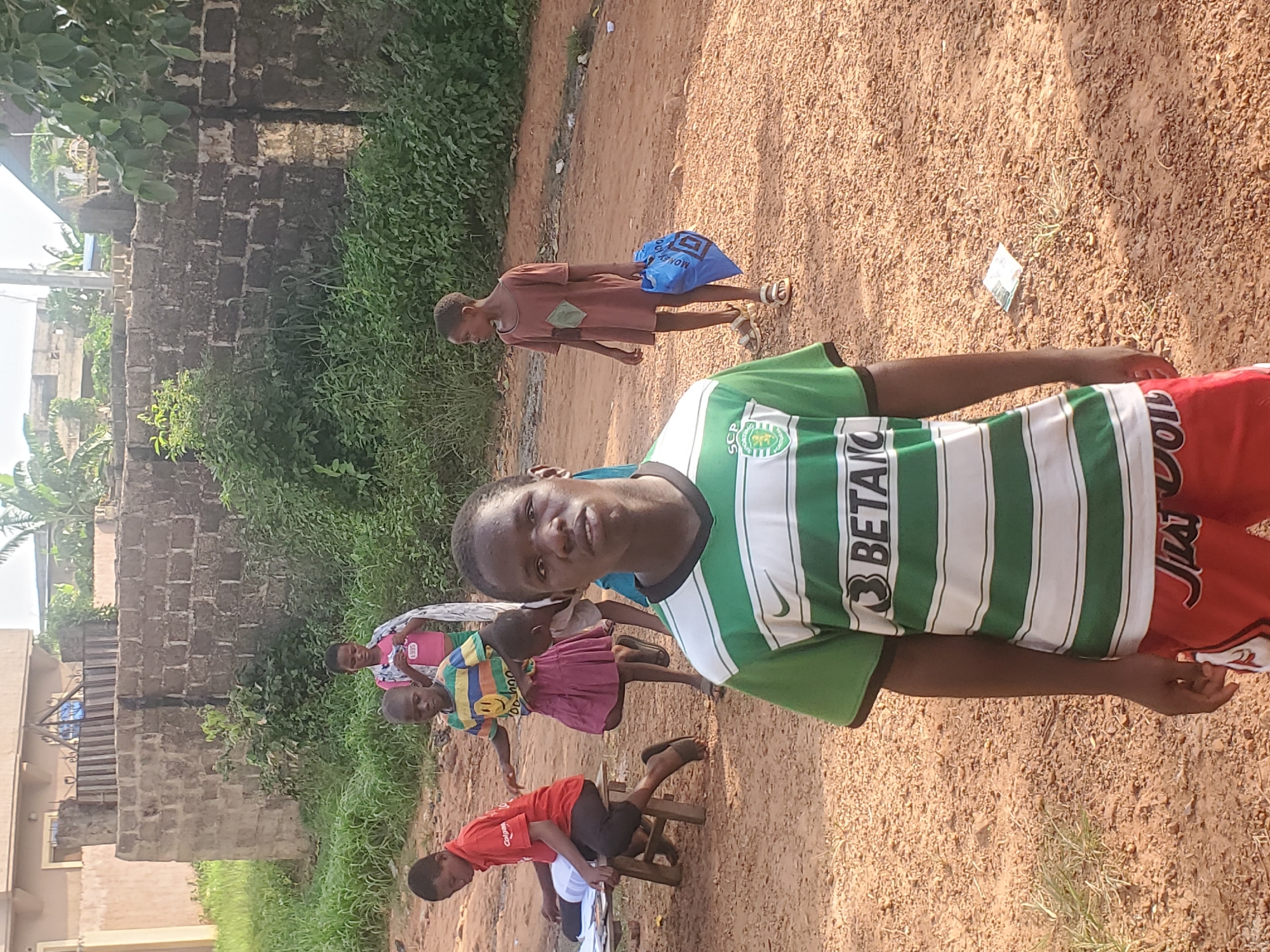 The image shows a group of children in an outdoor setting, likely in Ukwu-Abwa, Nigeria. In the foreground, a boy is standing and facing the camera, wearing a green and white striped jersey and red shorts. Behind him, several other children are visible. One child is sitting on a small wooden stool while wearing a red shirt. Another child is wearing a pink dress. In the background, a child walks away carrying a blue bag. The surroundings include a dirt ground, some vegetation, and a stone structure or wall. In the distance, there is a building.