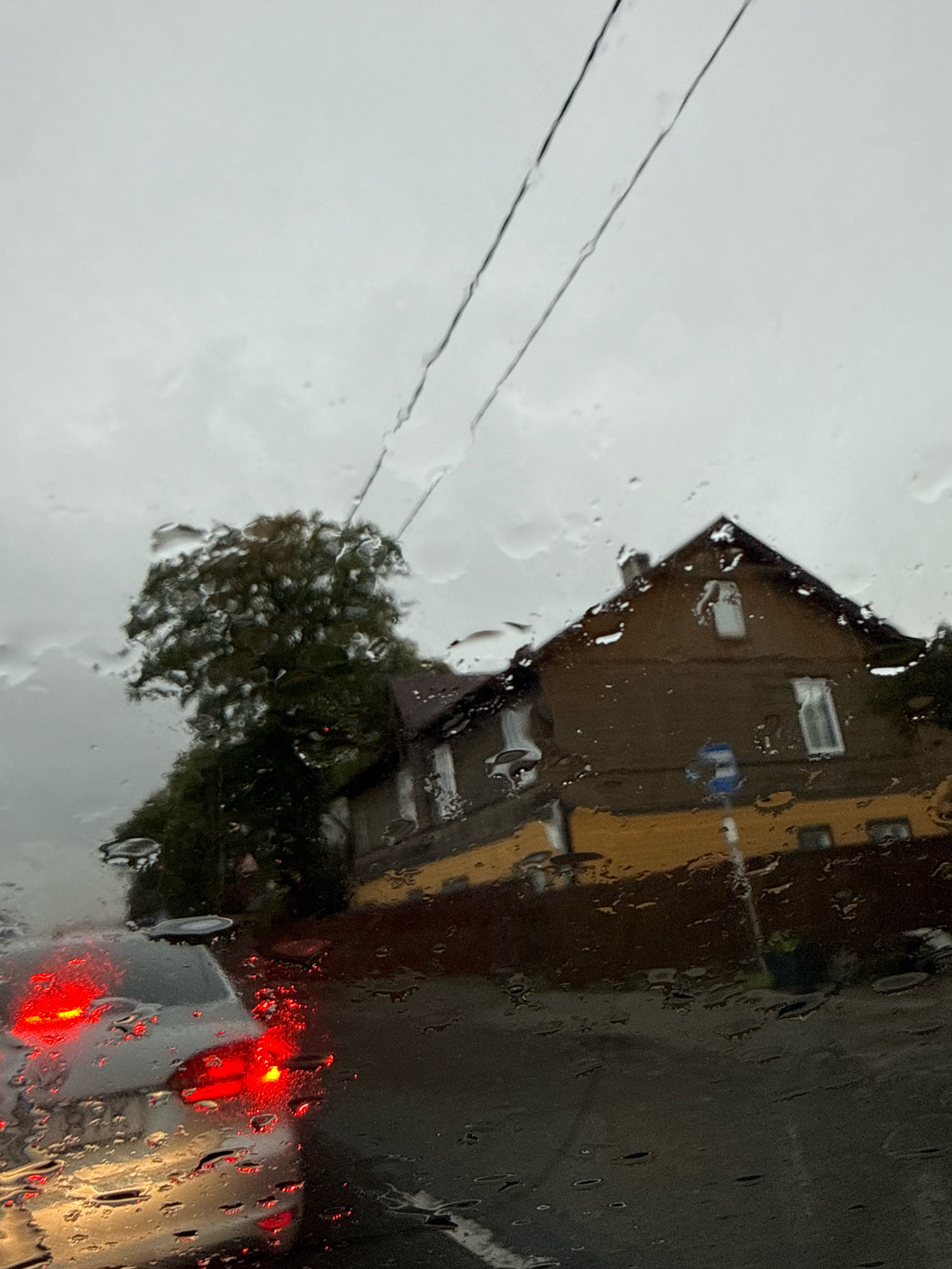 The image depicts a view through a rain-streaked car windshield, looking towards a street scene under an overcast sky. In the foreground, the tail lights of a car are illuminated, showing a red glow against a darker background with water droplets distorting the view. To the right, a two-story house with a dark brown upper story and a bright yellow lower story is visible. The house has several windows. A large tree with dark green foliage stands to the left of the house. Overhead, two dark utility wires run diagonally across the sky. A blue and white road sign is positioned near the house. The overall impression is a somewhat gloomy, wet day in a residential or mixed-use urban area, consistent with the city of Tallinn, Estonia.