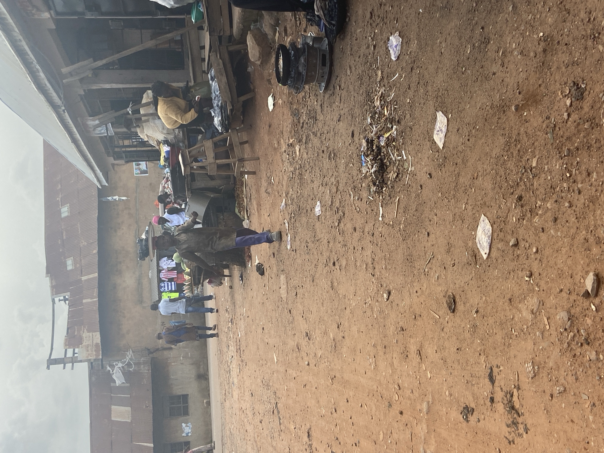 The image depicts a bustling street scene in Bukuru, Nigeria, with a focus on commercial activity and everyday life. The foreground is dominated by a dusty, red-brown dirt road, littered with various debris, including small stones, dried leaves, and pieces of paper or plastic. In the mid-ground, several individuals are engaged in commerce. A person in the center is walking, carrying a large load on their head, dressed in a dark, traditional garment. To their right, a stall displays colorful t-shirts, and next to it, another person is attending to goods on a wooden cart. Further to the right, a woman in a yellow top is bent over a collection of items on a wooden platform. In the background, a row of buildings with corrugated metal roofs suggests a market or a commercial area. The sky is overcast, contributing to a muted lighting condition. The overall impression is one of a vibrant, albeit somewhat unkempt, urban environment in Nigeria.