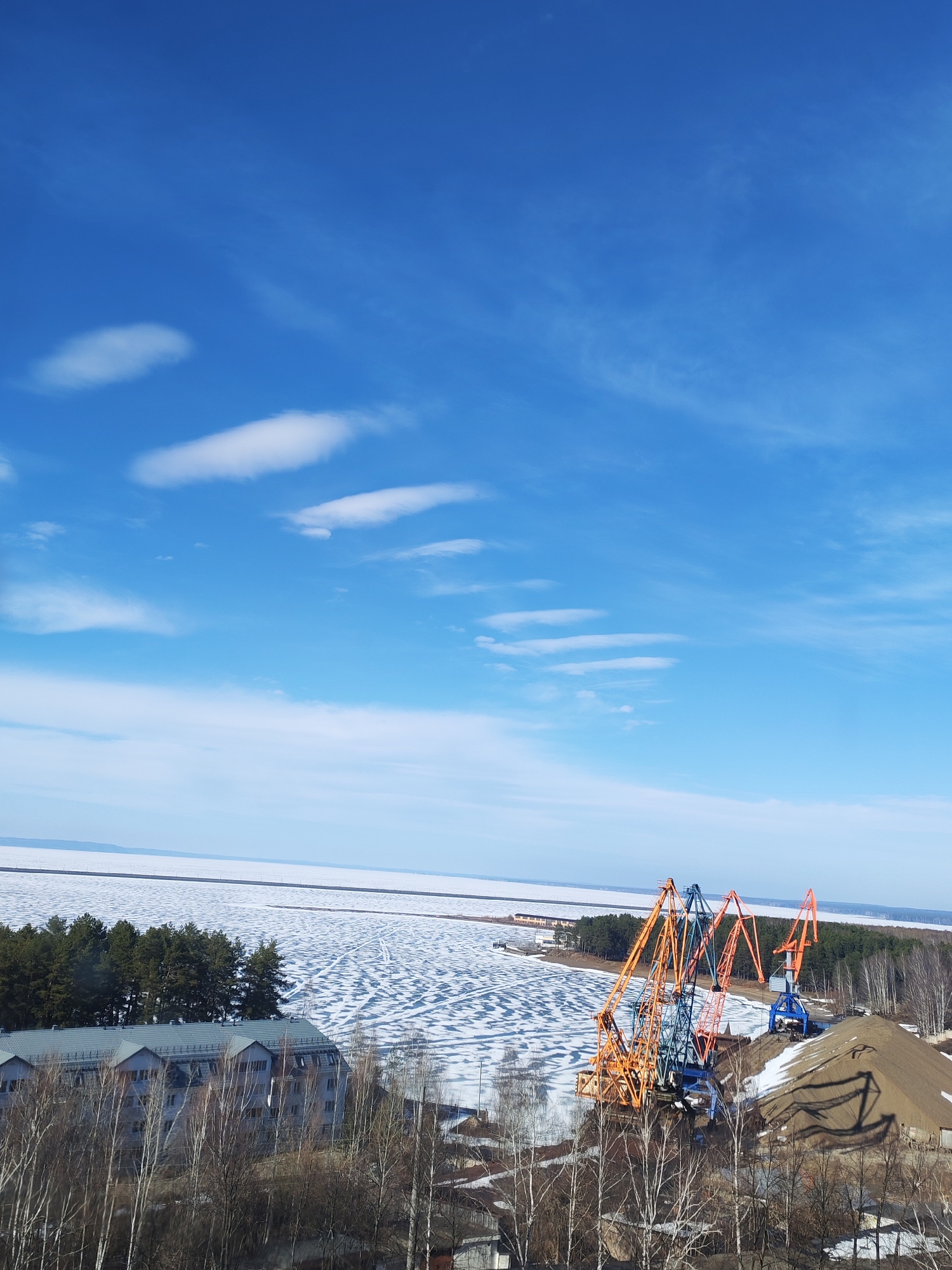 The image is a high-angle shot of a partially frozen body of water, likely the Kama River, near the city of Chaikovsky, Russia. The sky is bright blue with scattered white clouds. In the foreground, there are several industrial cranes, some orange and some blue, positioned near a large pile of sand or gravel. Behind the cranes, there is a forested area bordering the waterfront. Patches of ice cover the water's surface, with areas of open water visible. To the left, there is a multi-story building with a gray roof, nestled among bare trees. The overall scene suggests an industrial area near a river during a transition season, likely late winter or early spring.