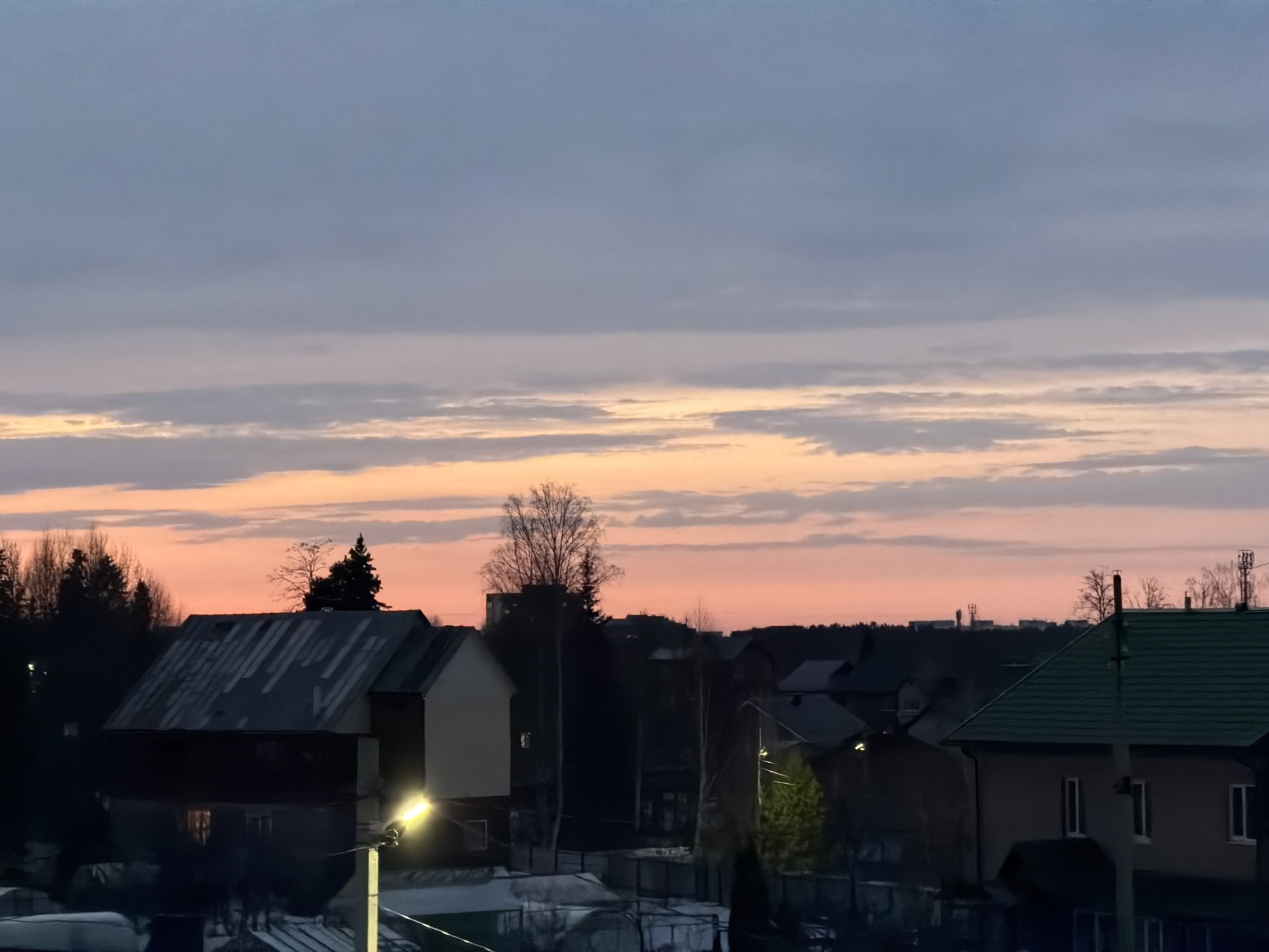 The image depicts a cityscape at dusk in Chaikovsky, Russia. The sky exhibits hues of gray, blue, and orange, indicative of a setting or rising sun. Residential buildings with various roof types, including slanted and green roofs, are visible in the foreground and midground. Trees, some bare and some with foliage, are interspersed among the buildings. Streetlights illuminate portions of the scene. In the background, silhouettes of buildings and trees line the horizon. The overall scene conveys a sense of tranquility and the onset of evening.