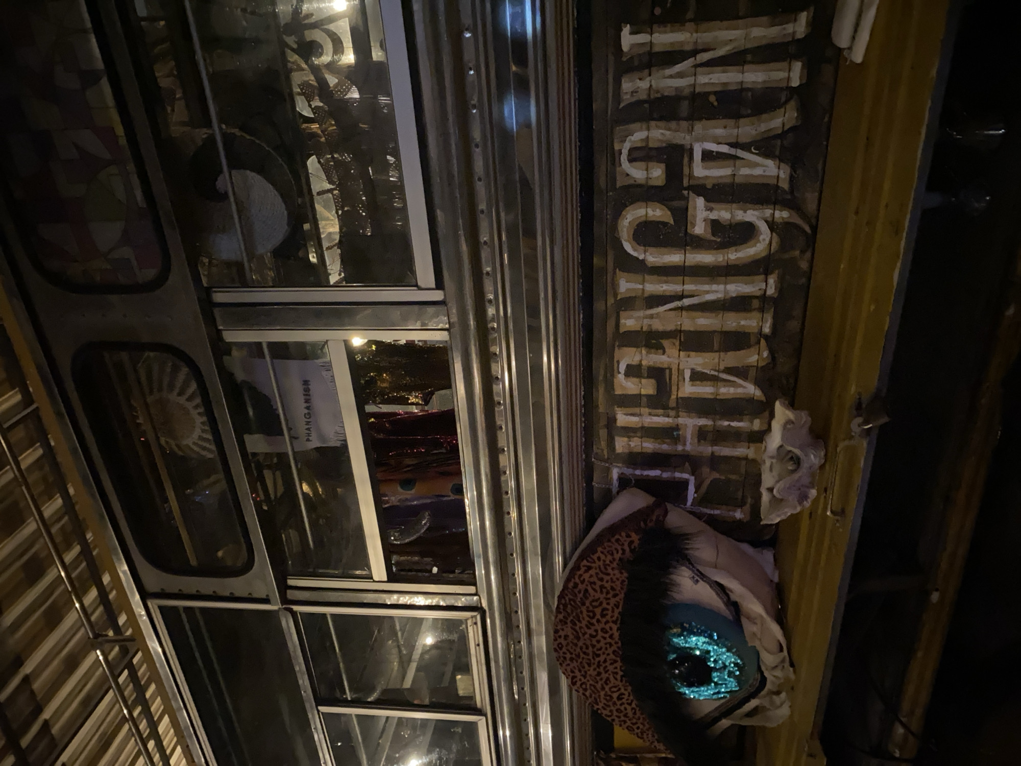 Captured at night in Baan Nai Suan, Thailand, the image shows the exterior of a decorative bus. The bus features multiple square windows, some reflecting interior elements. Its side displays vertically stacked lettering in white, reading "PHANGANISM." A decorative animal-print object and a white sculpted face are attached to the side of the bus. The environment appears dimly lit.
