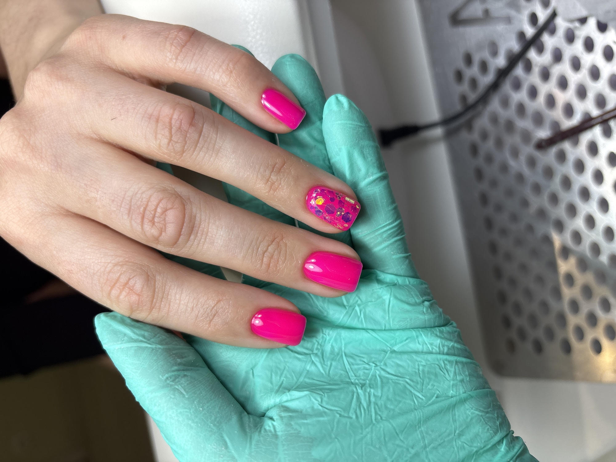 A close-up shows a hand with pink manicured nails, one of which has glitter. The hand rests on a gloved hand, likely of a nail technician, inside a salon. The background shows part of nail salon equipment. It is not possible to confirm the location in Ферма, Russia from the given image.