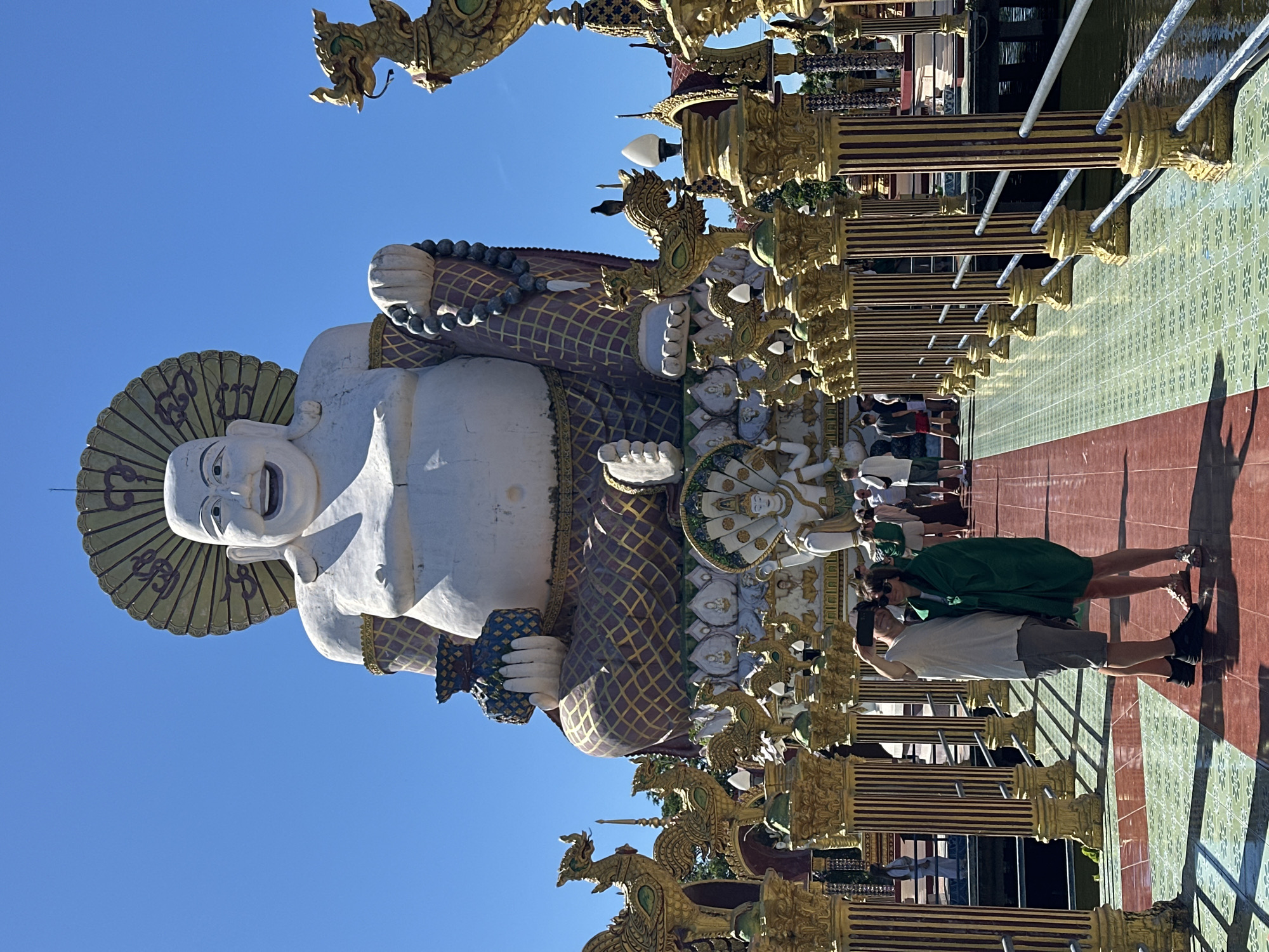 This image captures a scene at the Big Buddha Temple (Wat Plai Laem) in Baan Plai Laem, Thailand. Dominating the background is a colossal white statue of a laughing Buddha, seated and adorned with a patterned robe and beaded necklace. The Buddha statue is framed by a large, fan-like halo. In the foreground, a pathway with red and green tiled flooring leads towards the temple. Flanking the pathway are rows of ornate, golden pillars topped with decorative dragon-like figures and lamps. A couple is positioned on the pathway, facing away from the camera, with one person taking a photo of themselves with a smartphone. Other visitors are visible further down the pathway and in the background, amidst the temple's elaborate architecture. The sky above is clear blue, indicating a sunny day.