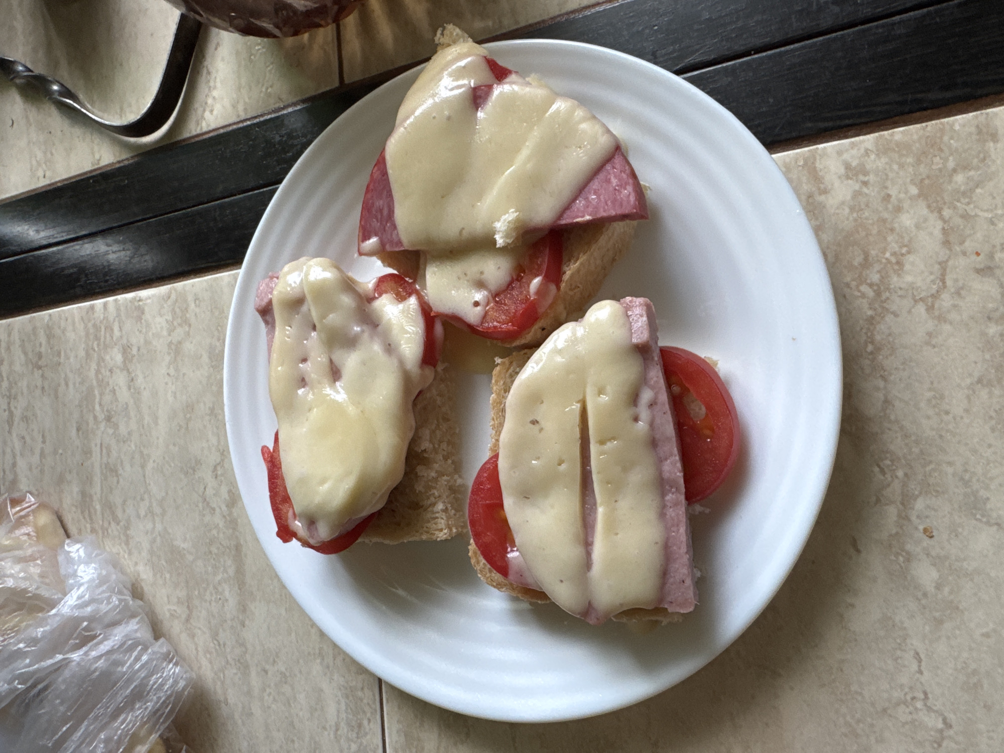Three open-faced sandwiches are arranged on a white plate. Each sandwich consists of a slice of bread topped with a slice of tomato, a slice of ham, and a slice of melted cheese. The sandwiches are placed on a tiled surface. In the background, a plastic bag is visible. The overall scene appears to be an indoor setting, possibly a kitchen or dining area, in Комарихинский, Russia.