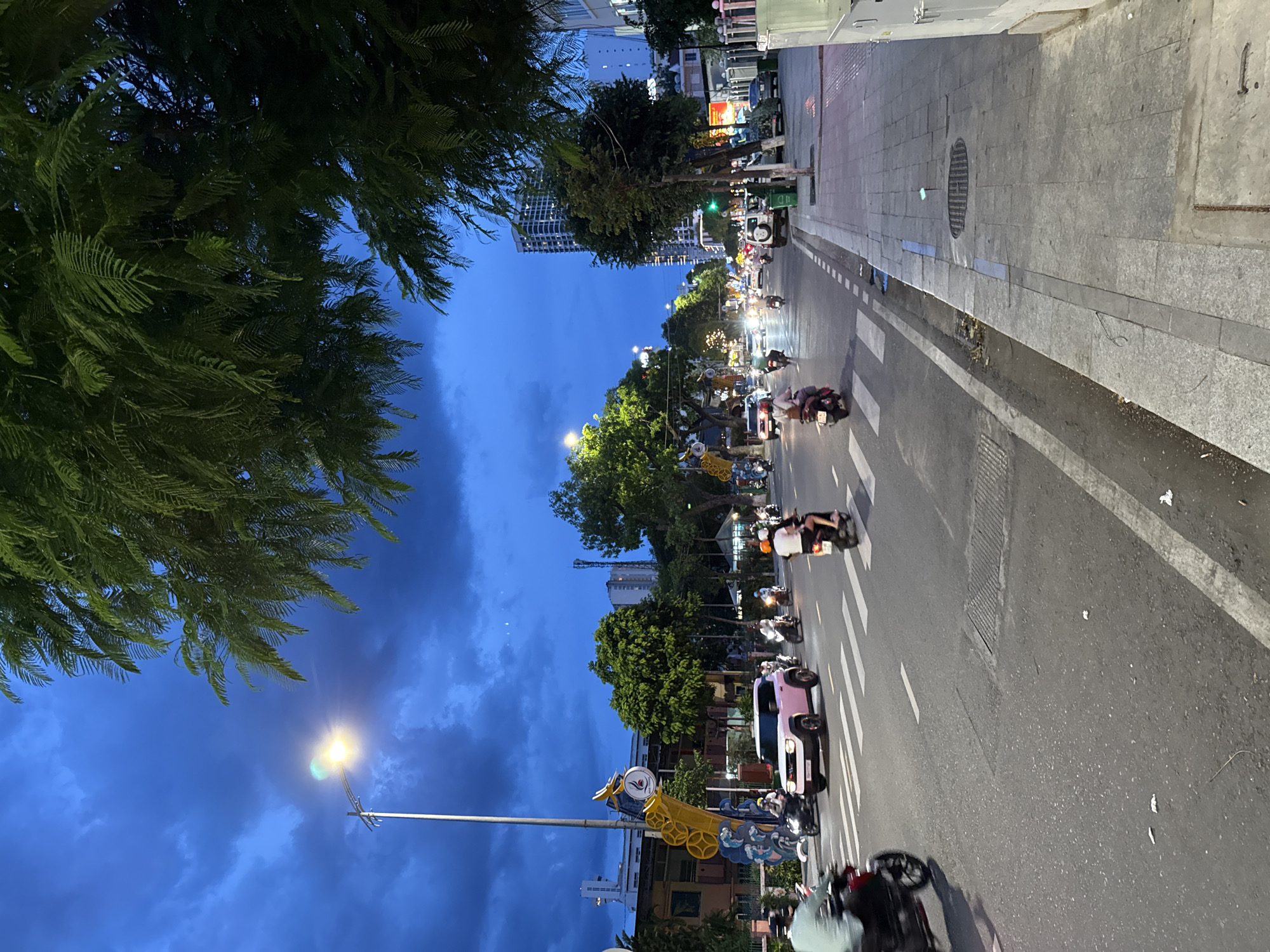 The image depicts a street scene in Nha Trang, Vietnam, during twilight. The sky is a deep blue with dramatic clouds, illuminated by streetlights and the headlights of vehicles. A large tree with dark green foliage dominates the upper portion of the frame, casting shadows over the scene.
The street is busy with traffic, primarily motorcycles, but also includes cars and what appears to be a pink SUV. People are seen riding on the motorcycles, some wearing helmets. Pedestrians are visible on the sidewalks, which are paved with stone tiles. Buildings line the street, including a tall modern high-rise in the background.
The overall impression is of a vibrant urban environment transitioning into evening, with elements of both natural and man-made illumination. The scene suggests a typical street in a Vietnamese city, characterized by lively traffic and a mix of architecture.