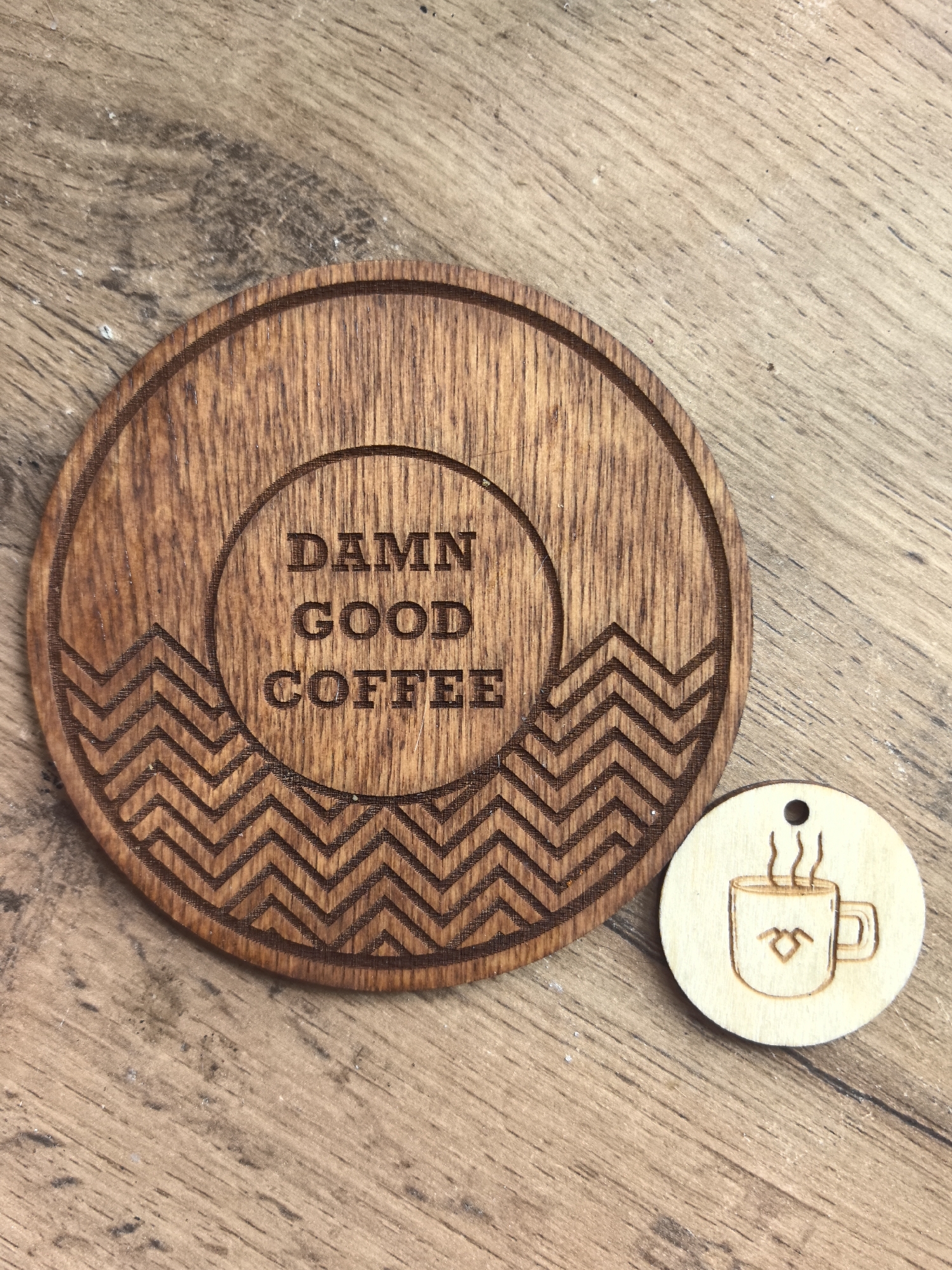 The scene is located in the city of Noviy, Russia. The image displays two distinct wooden items positioned on a light brown wooden surface with visible grain and minor surface imperfections. A large, circular wooden coaster, dark brown in color, occupies the central left portion of the frame. It features engraved text in the center reading "DAMN GOOD COFFEE" arranged in three lines within a circular border. Below this text, a concentric band of engraved chevron patterns is present. To the lower right of the coaster rests a smaller, lighter-toned circular wooden tag. This tag has a small hole at its top edge and features a simple engraved illustration of a steaming coffee mug, which includes a heart-like or stylized shape on its side. No individuals or dynamic actions are depicted.