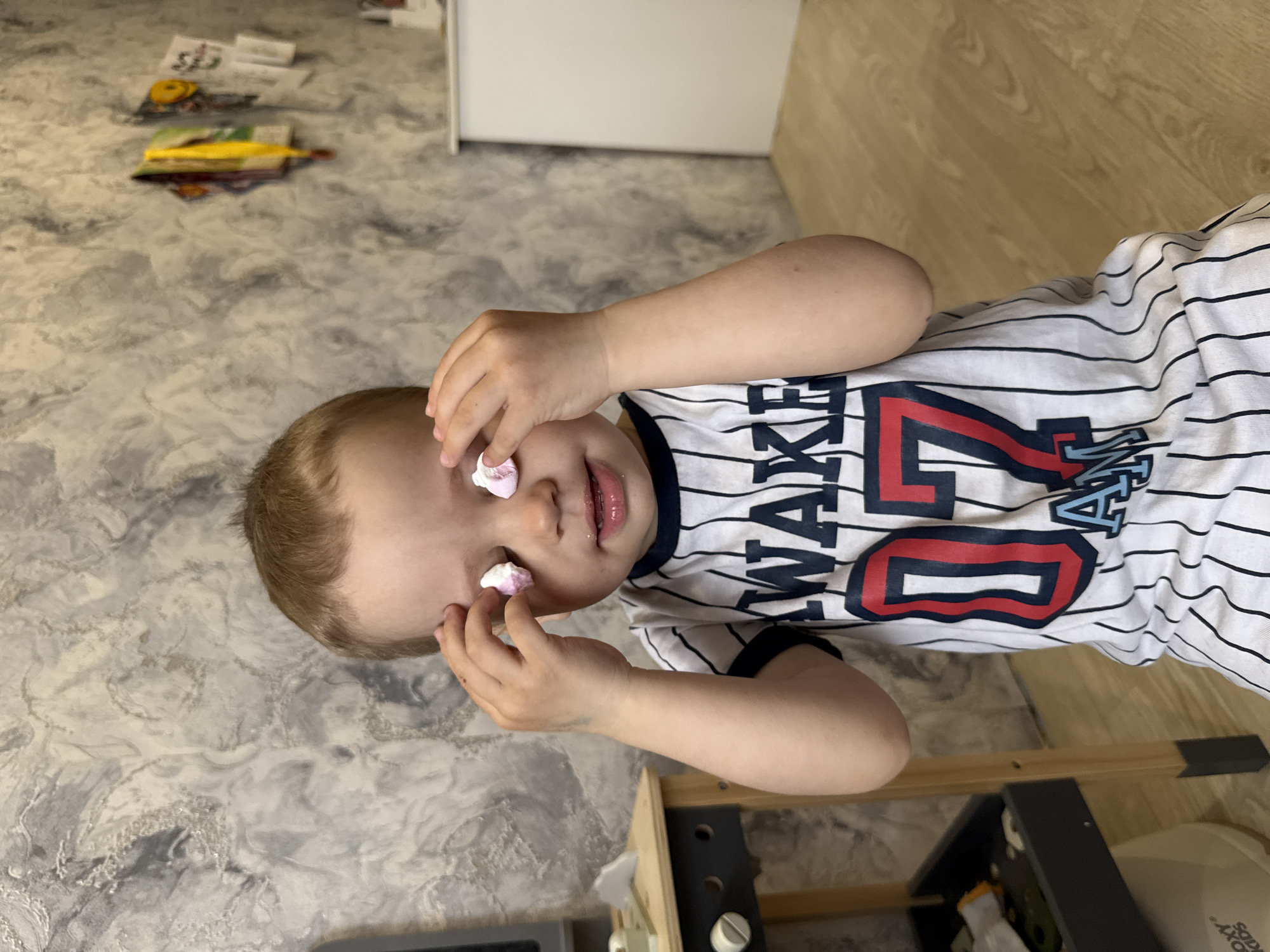 A young child, appearing to be a toddler or preschooler, is depicted indoors in a playful moment. The image is oriented sideways, showing the child's upper body and face. The child has fair skin, short light brown hair, and rosy lips, slightly parted in a faint smile, indicating happiness or amusement. They are wearing a white t-shirt with thin black horizontal stripes. Prominently displayed on the shirt is the word "WAKE" in large black letters, followed by a large red number "20" (or similar numbers) with a black outline, and other blue letters that resemble "LA" or "NY", giving the shirt a sports jersey aesthetic.
The central activity involves two small, light-colored confectioneries, possibly meringues or marshmallows (one appearing white, the other slightly pink), placed on each of the child's closed eyes. One of the child's hands is raised near their face, as if just having placed or adjusting the sweets.
The setting appears to be a home in Rusanovo, Russia. The floor features a distinct grey and white swirling pattern, transitioning to a light wood-look floor on the right side of the frame. In the background, to the upper left, there are some items scattered on the patterned floor, including papers, what looks like books, and a yellow object. Part of a white cabinet or wall is visible behind these items. To the lower right, a wooden furniture frame and a dark grey or black furniture piece are partially visible. Text "KIDS" is faintly visible on a white object near the bottom right corner of the frame. The overall lighting is bright and even, suggesting an indoor daytime scene.