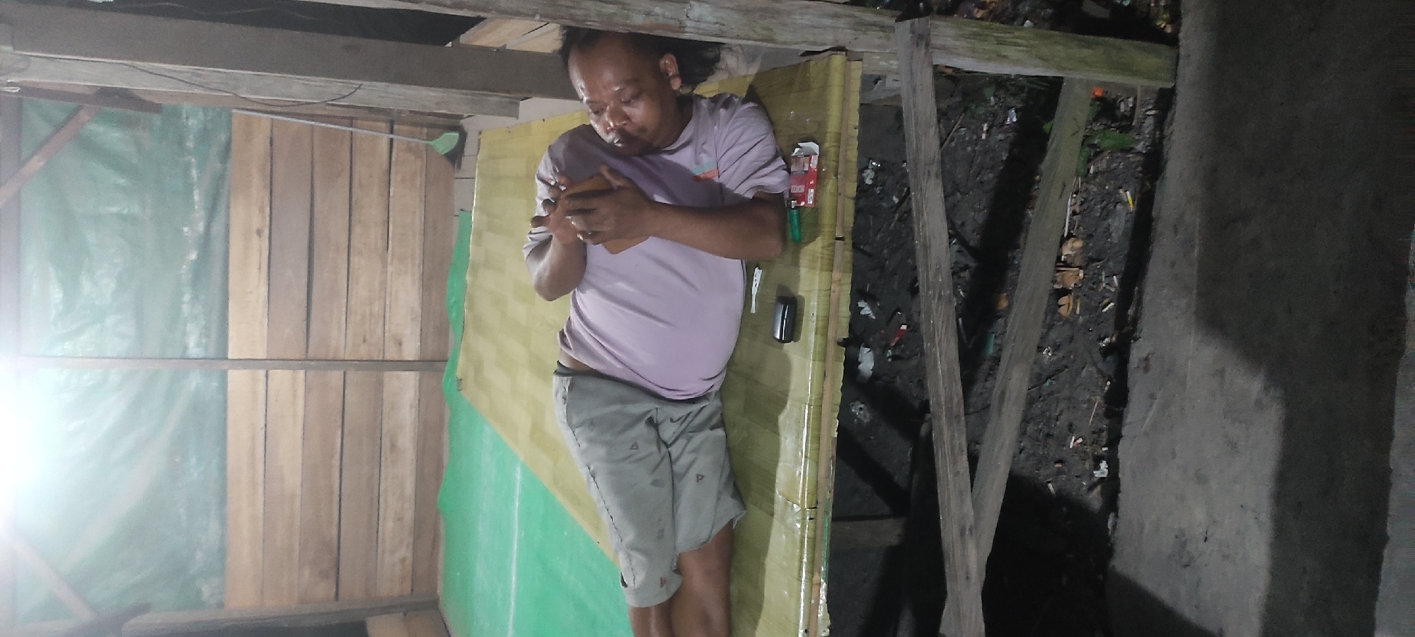 A man is lying on his right side on a raised wooden platform, covered with a yellow mat. He is wearing a light purple t-shirt and grey shorts. The man is holding a mobile phone in his hands and appears to be using it. To his right, on the mat, are a black rectangular object, possibly a power bank, a cigarette lighter, and a partially open red cigarette pack. The platform is situated in an open-air structure with wooden plank walls in the background, partially covered by a green tarp. Beneath the platform, the ground is dirt with scattered debris, including cigarette butts and wood fragments. The scene is dimly lit, suggesting it is either evening or night. The location is identified as Bumi Harjo, Indonesia.