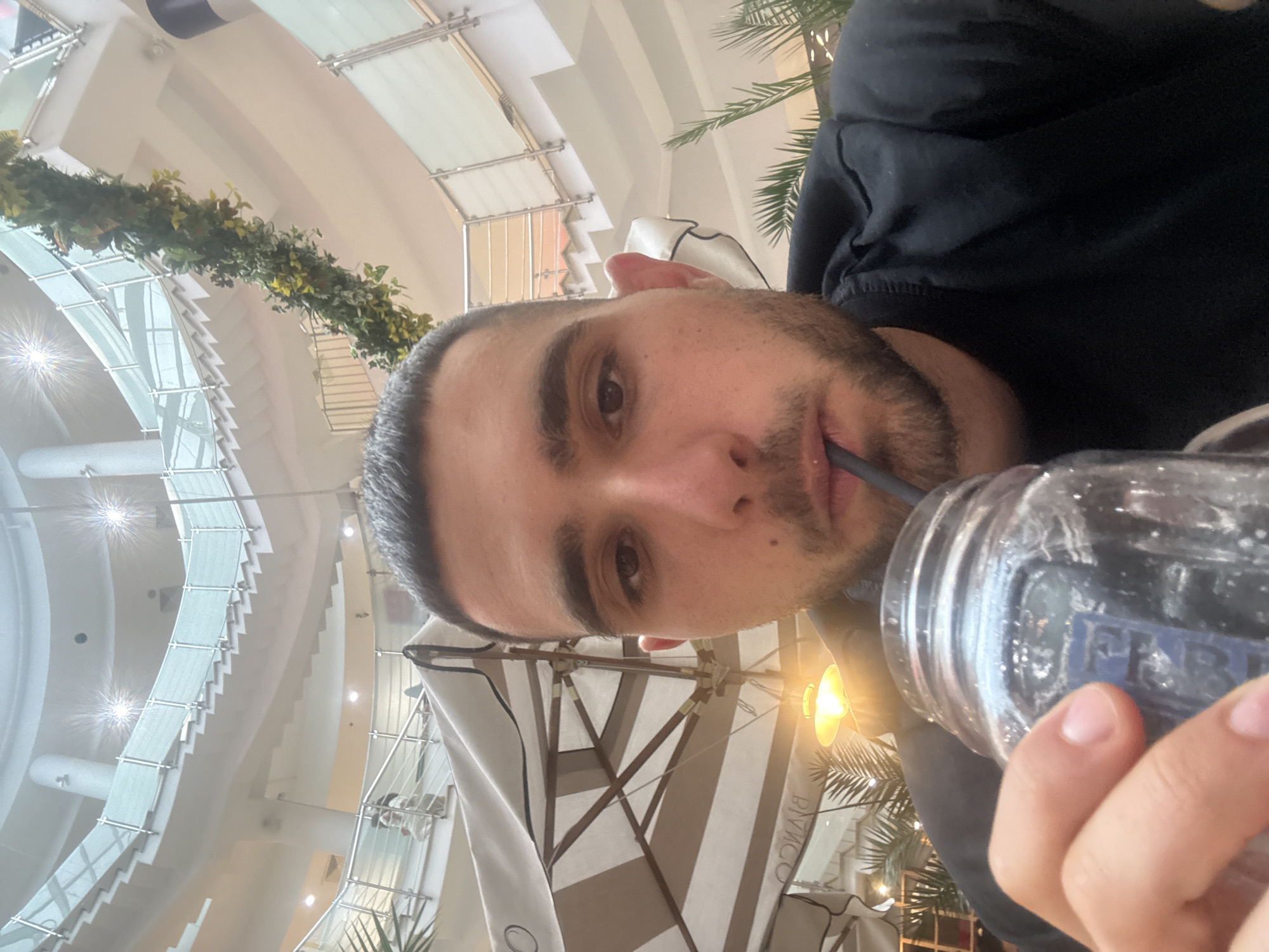 A male individual, estimated to be between 25 and 35 years old, with short dark hair and a beard, is depicted in a close-up, upward-angled photograph, consuming a beverage through a black straw from a clear glass mason jar held in his right hand. He is wearing a dark-colored, possibly black, long-sleeved top or hoodie.
The background reveals a multi-level, brightly lit interior architectural space, identified as being in Veliko Tarnovo, Bulgaria. The structure features sweeping, curved white staircases with glass and metal railings that ascend through an open atrium. Several bright, starburst-effect light fixtures are visible on the upper levels. A dense garland of green and yellow artificial foliage is draped along an upper railing on the right side of the frame.
To the left of the subject, a large, partially open outdoor-style umbrella with beige and white stripes is present, displaying the reversed text "BINGO". Below the umbrella, a warm, bell-shaped light fixture illuminates the area. To the right of the subject, dark green, palm-like artificial plants are visible. Faint figures of other individuals can be observed on the upper levels of the building. The overall environment suggests a cafe or dining area within a large commercial complex, likely a shopping mall, in Veliko Tarnovo.