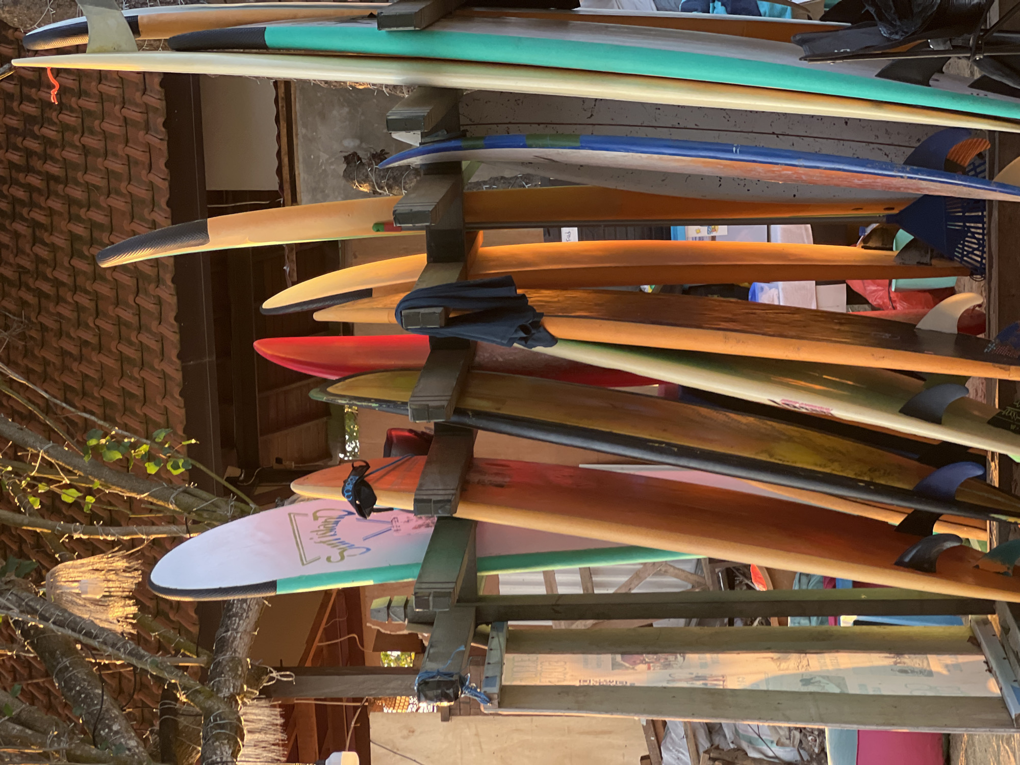 A collection of surfboards is displayed vertically in a wooden rack. The surfboards are of various colors and designs. Some have graphics on them, while others are plain. A dark blue cloth is hanging from one of the surfboards. The background shows a tiled roof and trees. This scene is located in Kuta, Bali, Indonesia, known for its popular beaches and surf culture.