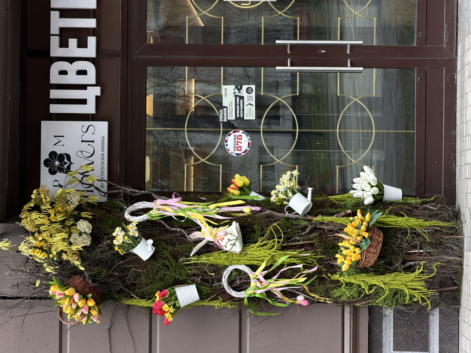 A storefront display in Khimki, Russia, features a brown-framed glass door with gold geometric designs. Signs and stickers are affixed to the glass, advertising services and job openings at "Moné". The entrance is decorated with a large arrangement of flowers, moss, and branches, incorporating elements like watering cans and small baskets. The display includes white lettering, part of which reads "ЦВЕТ", and a sign that reads "M Flowers BY PETROVSKAYA MARINA".