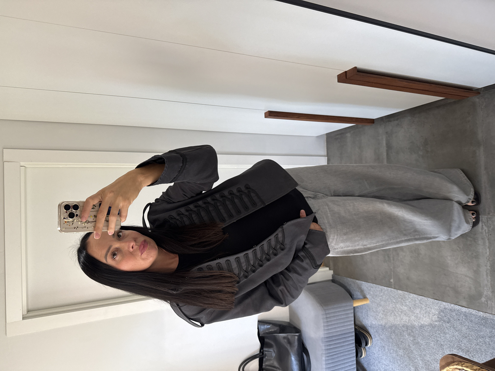 A woman with long, dark hair is taking a mirror selfie indoors. She is wearing a dark gray military-style jacket with ornate braiding and a black t-shirt underneath, paired with loose-fitting light gray jeans. Her right hand holds a smartphone with a floral-patterned case, and her left hand is in her pocket. The background shows a white wall with vertical paneling, a full-length mirror reflecting the scene, and a doorway to the left. To the left of the woman, there is a gray upholstered bench with a black leather tote bag resting on it, and a pair of black sandals visible underneath. The floor is a mix of concrete and gray carpet. The overall setting appears to be a domestic interior, possibly a dressing room or hallway. The image does not contain any elements that suggest a location in Chaikovsky, Russia.