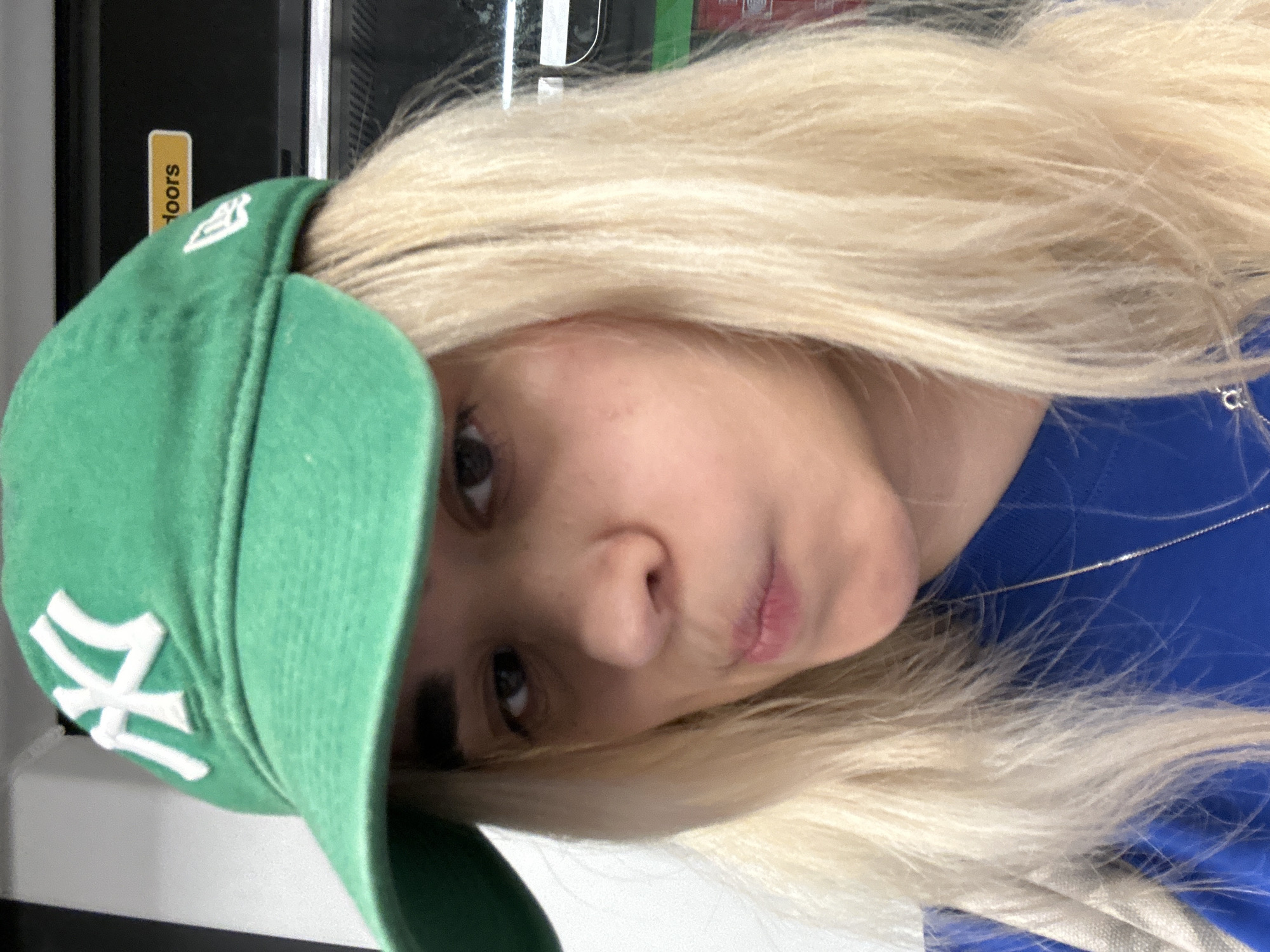 A close-up portrait depicts a person with long, blonde hair, wearing a green baseball cap and a blue top. The subject's head is tilted slightly, and they look towards the viewer with a neutral or subtly pursed-lip expression.
The bright green baseball cap is worn on the head, featuring a prominent white "NY" logo on the front, characteristic of the New York Yankees. A smaller white flag-shaped logo, likely belonging to New Era, is visible on the left side of the cap. The person's long, wavy blonde hair cascades around their face and shoulders. A thin silver chain necklace is visible against the blue fabric of their top.
The background appears to be indoors, consisting of dark, possibly reflective surfaces and a vertical yellow sign. Partially visible text on this sign reads "doors". The lighting is soft and even, suggesting an indoor setting.