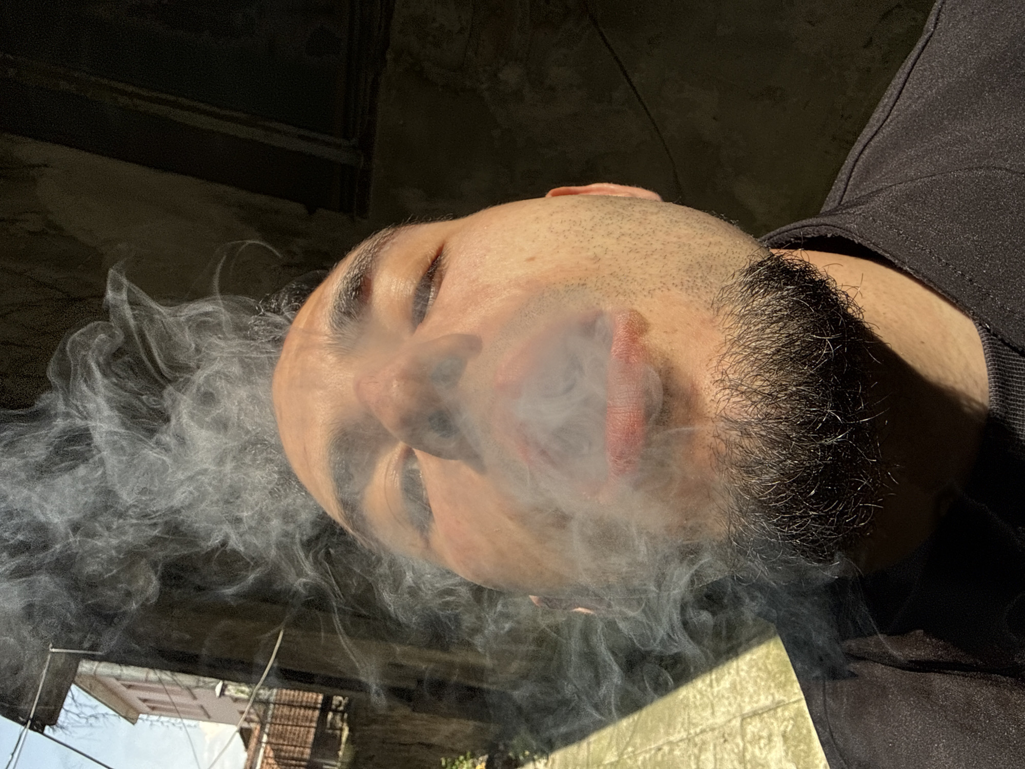 A man exhales smoke in an outdoor setting. He has dark hair, a beard, and is wearing a black shirt. The background includes a building and some greenery. The specific location is difficult to determine with certainty.