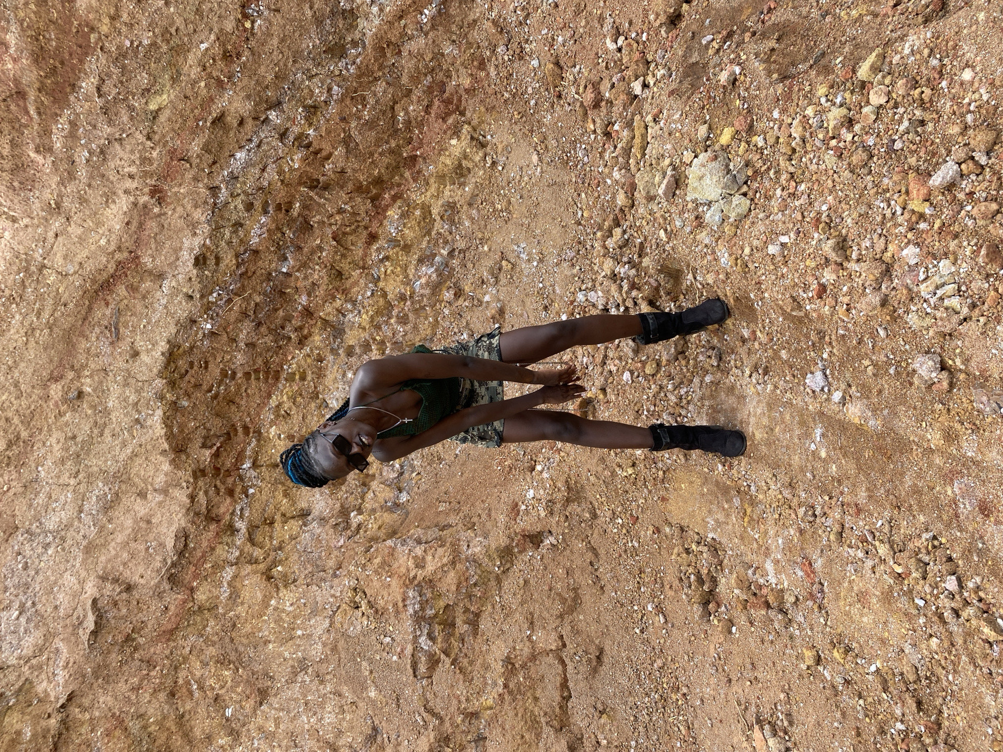 The image shows a young Black woman standing in an outdoor environment characterized by rough, reddish-brown terrain. The ground is covered with loose soil and various sized rocks, suggesting a natural or excavated area. Behind her, a steep, layered rock formation dominates the background. The woman is wearing a dark green, possibly camouflaged, skirt and a matching top. She has dark sunglasses on and her hair is styled in blue braids. She is posing for the camera with her hands in front of her. The overall scene appears to be a natural, undeveloped area. There is no specific visual information in the image that can confirm or deny its location in Bar Jirgi Summa, Nigeria.