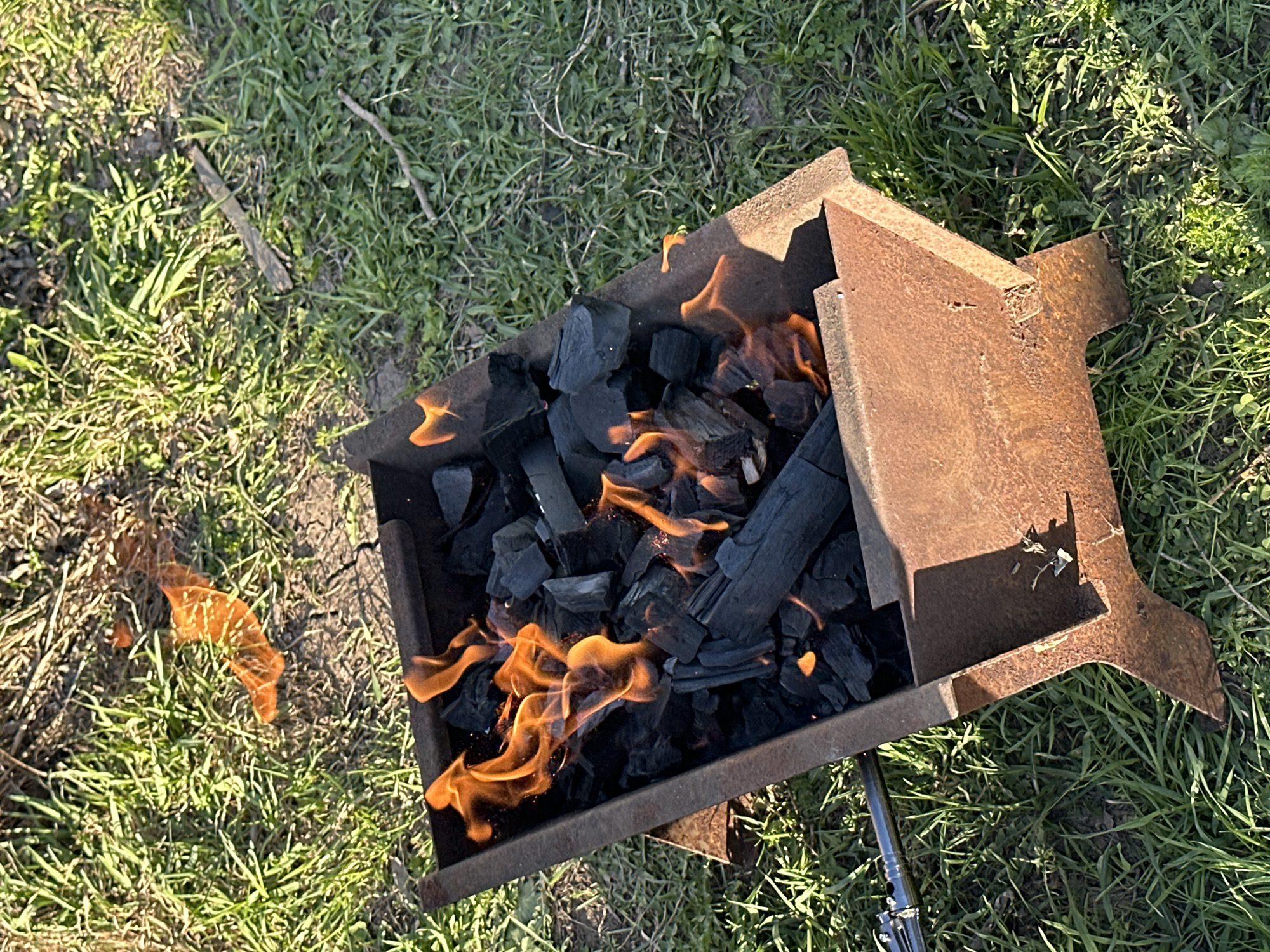 A high-angle outdoor shot shows a rusty metal barbecue pit containing burning charcoal. Flames are visible among the charcoal within the pit and also extend from the pit onto the surrounding grass. The barbecue pit has a simple square design with crude, cutout legs. The environment is grassy with patchy areas of bare soil and some fallen twigs. The location is Среднее Кечево, Russia. No people are visible.