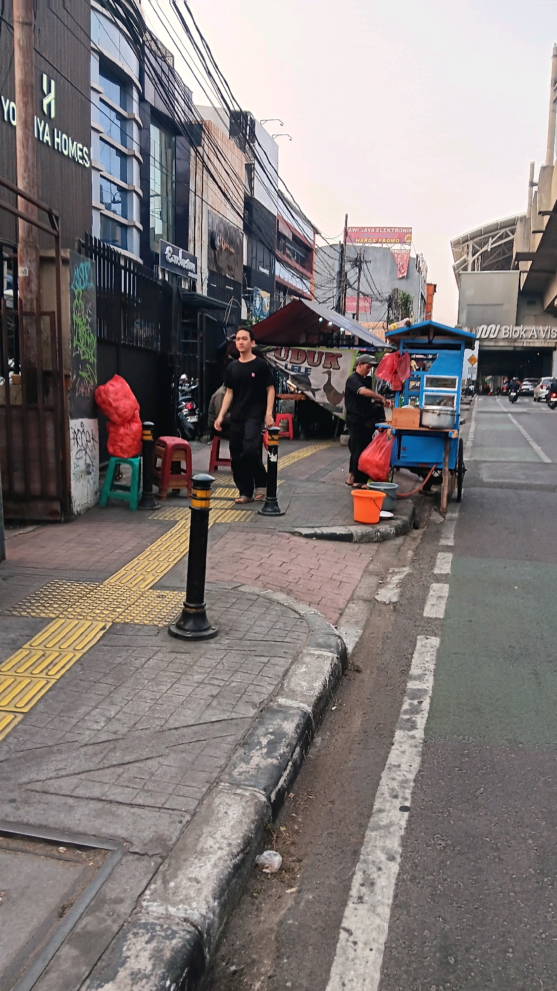 The image shows a street scene in South Jakarta, Indonesia, likely during the late afternoon or early evening given the lighting. In the foreground, a sidewalk with tactile paving for the visually impaired runs alongside a road. A black bollard with yellow markings is positioned on the sidewalk. Two people are present: one person in a black t-shirt and pants stands on the sidewalk, appearing to walk towards the viewer. Further down the sidewalk, another person in a black t-shirt and pants is standing behind a blue food cart, interacting with it.
The food cart is blue and appears to be selling food. It has shelves displaying items and a canopy over the top. Next to the cart, an orange bucket and a grey bucket are visible on the ground. In the background, there are several buildings. The building on the left has signage that reads "YOI YA HOMES". Further down the street, more buildings are visible, including one with a red sign advertising "AWI JAYA ELEKTRONIK HARGA PROMO". A pedestrian overpass or elevated structure is visible in the far background. The environment appears to be a busy urban area with overhead power lines and some street vegetation.