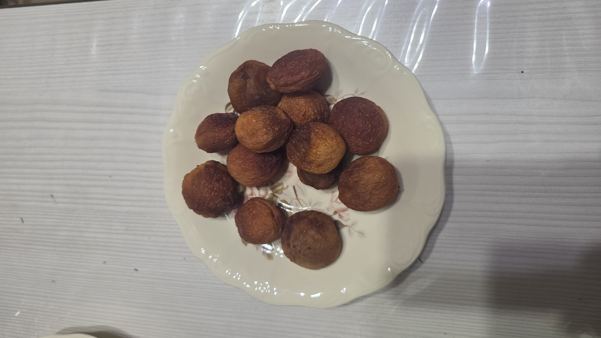 The image shows a white, scalloped-edge plate with a floral pattern, filled with dried apricots. The plate rests on a surface with a light-colored, vertical striped pattern, possibly a tablecloth or a textured placemat. The apricots are a deep orange to reddish-brown color, indicating they are dried. There are approximately twelve dried apricots visible on the plate, arranged in a somewhat scattered manner. The lighting is moderate, casting soft shadows. There are no people or other objects clearly identifiable in the scene, nor is there any indication of a specific location in Chuy, Kyrgyzstan. The focus is solely on the plate of dried fruit.