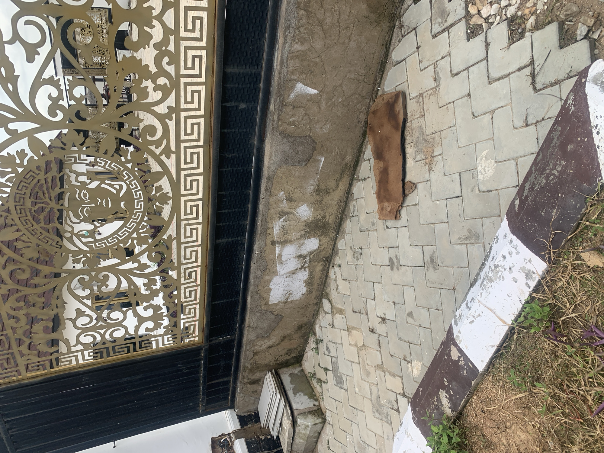 The image shows a close-up of an ornate metal gate, likely an entrance to a property, in Jauro Yinu, Nigeria. The gate features a decorative pattern with a Greek key motif and a stylized human face, possibly Medusa, at its center. The gate is supported by black metal posts and is situated above a concrete wall with visible weathering and some white paint marks.
In front of the wall is a paved walkway made of interlocking grey square tiles arranged in a herringbone pattern. The walkway has a curb on the left side, painted with alternating white and dark brown stripes, showing signs of wear and tear. A patch of dry grass and some green plants are visible near the curb.
On the paved walkway, in the foreground and to the right of center, lies a flat, irregularly shaped piece of brown material, possibly a piece of discarded wood or roofing tile. To the left of this object, a stack of flat grey rectangular objects, resembling tiles or slates, rests on a concrete base. Small stones and debris are scattered along the edge of the paved area on the right side of the image. The overall scene suggests an outdoor environment, possibly a residential or public area, with elements of construction or disrepair.