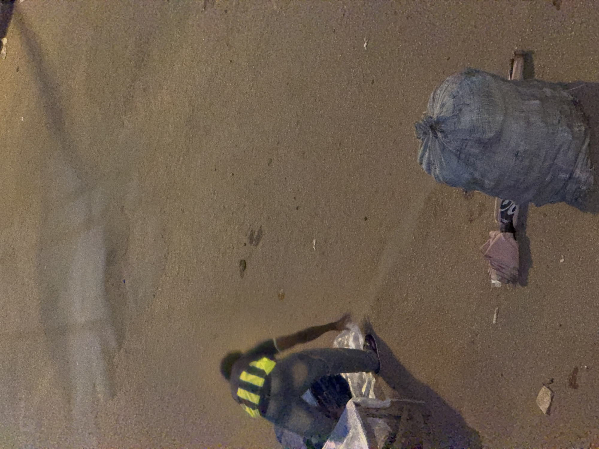 In Bukuru, Nigeria, a person wearing a dark shirt with yellow stripes and blue jeans is bent over on a paved surface, reaching towards a white object on the ground. To their left, there is a white structure with some objects on top, and more fabric or bags are piled near the person's legs. A large, grey, duffel-like bag is situated to the right of the frame, and other smaller items, including a folded pink material, are scattered on the ground between the person and the large bag. The environment appears to be an outdoor, urban setting, likely a street or open area with rough paving.