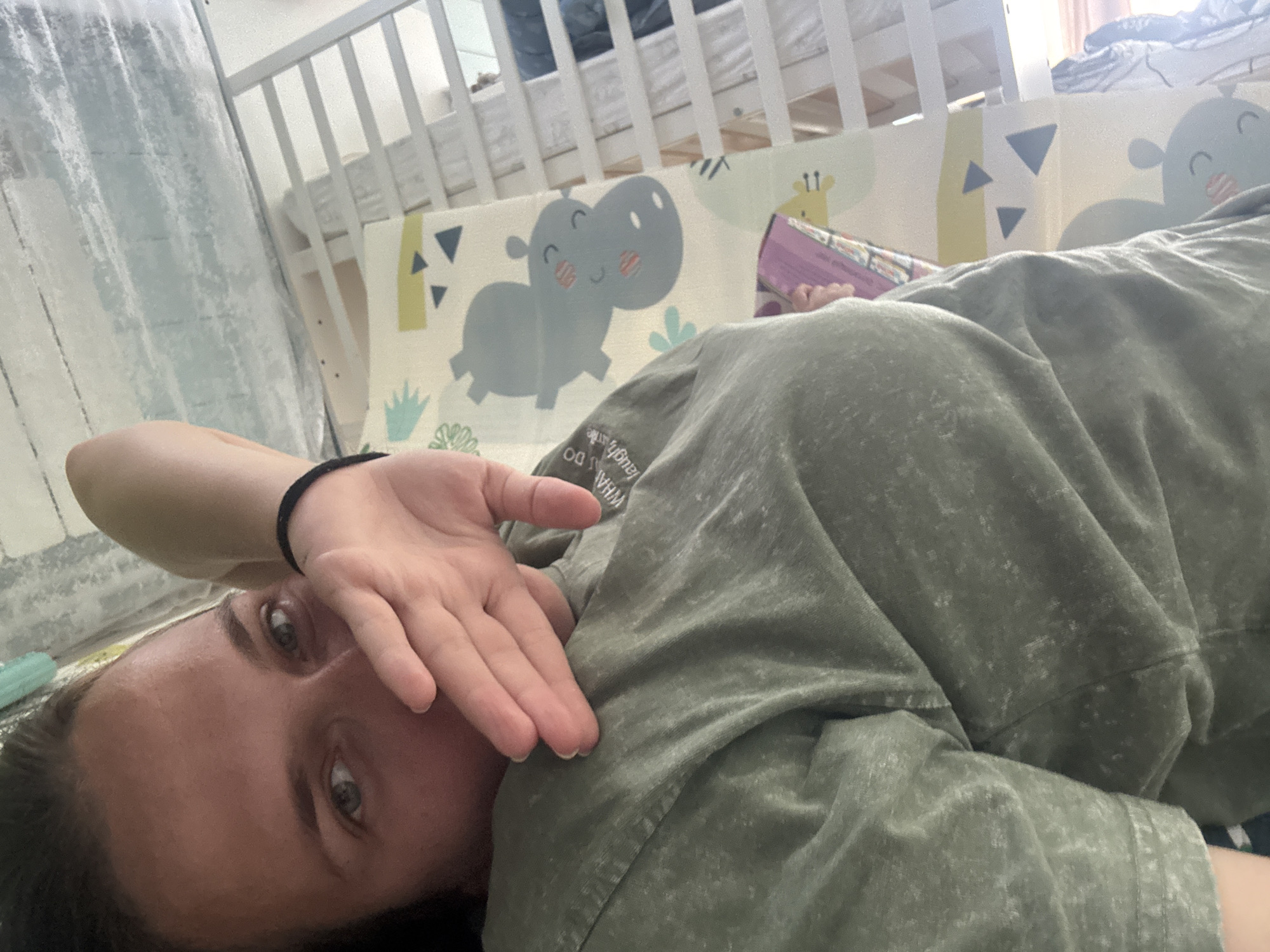 The image captures a close-up, angled view of a person lying down indoors. The subject, seemingly female, looks directly into the camera, with her left hand partially covering her face. A black elastic hair tie is visible on her left wrist. She is wearing a green or khaki t-shirt with faded white text, which partially reads "WHAT DO you LAUGH at me".
In the background, to the left, a light-colored curtain or fabric hangs. To the right, a white wooden crib is visible, and in front of it, a children's playmat or bumper pad displays cheerful cartoon animals, including a grey hippopotamus with rosy cheeks, yellow giraffe spots, and geometric triangle patterns. A small hand, presumably a child's, can be seen on the right, holding a book with a pink cover, resting near the playmat. The scene suggests a casual domestic setting, possibly a child's room or play area in Razdolye, Russia, during daylight hours. The person appears to be resting or observing, potentially feeling tired or relaxed in the vicinity of a child.