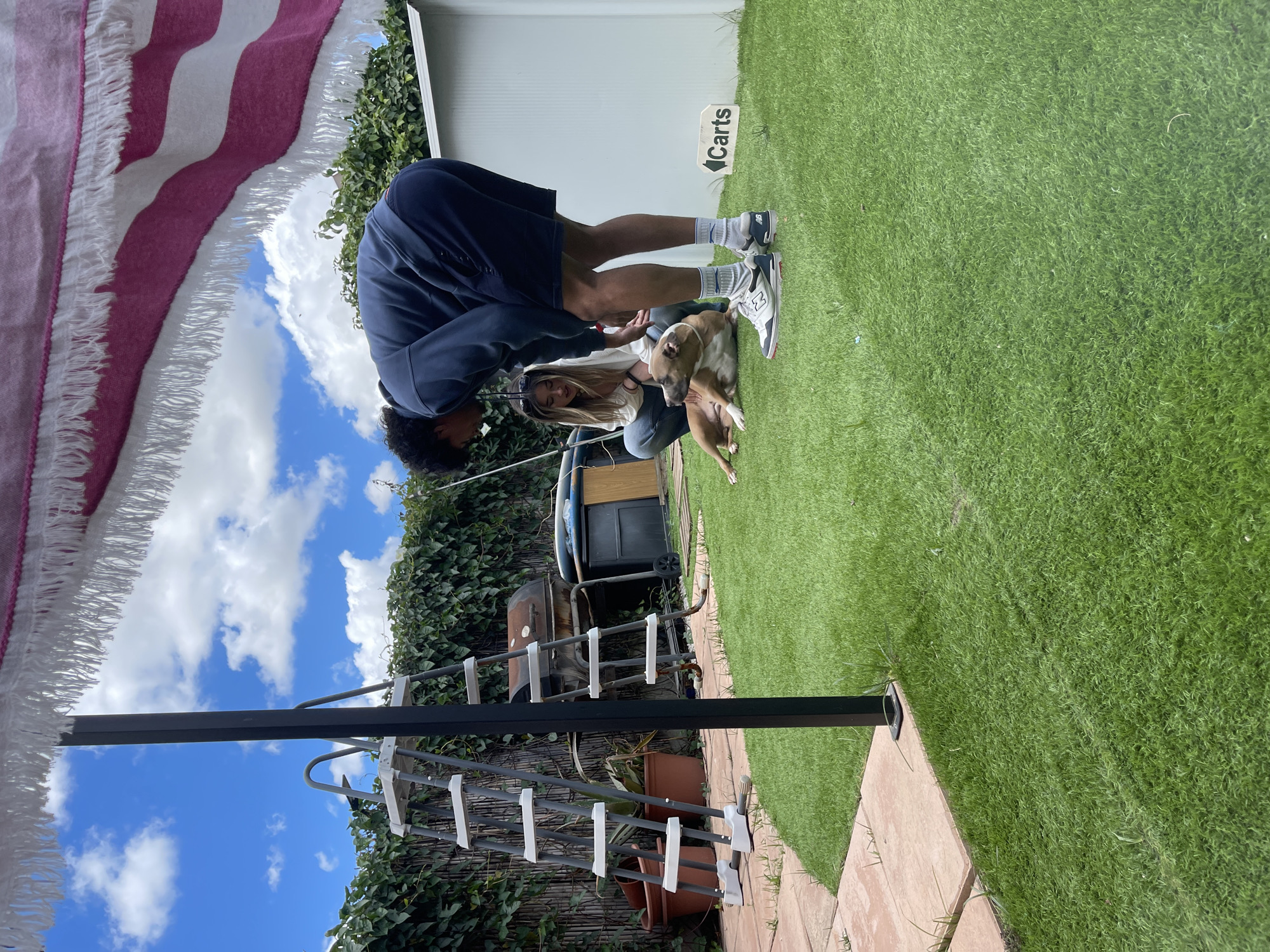 Outside in a fenced yard in Jerez De La Frontera, Spain, a man and woman are tending to a dog on the grass. The man, wearing dark shorts, a dark sweatshirt, and sneakers, is bending over the dog. The woman, wearing jeans and a white top, is kneeling next to the dog. Behind them is a white building with a "Carts" sign. A grill, planters, and a towel rack are also present. The sky is blue with clouds.